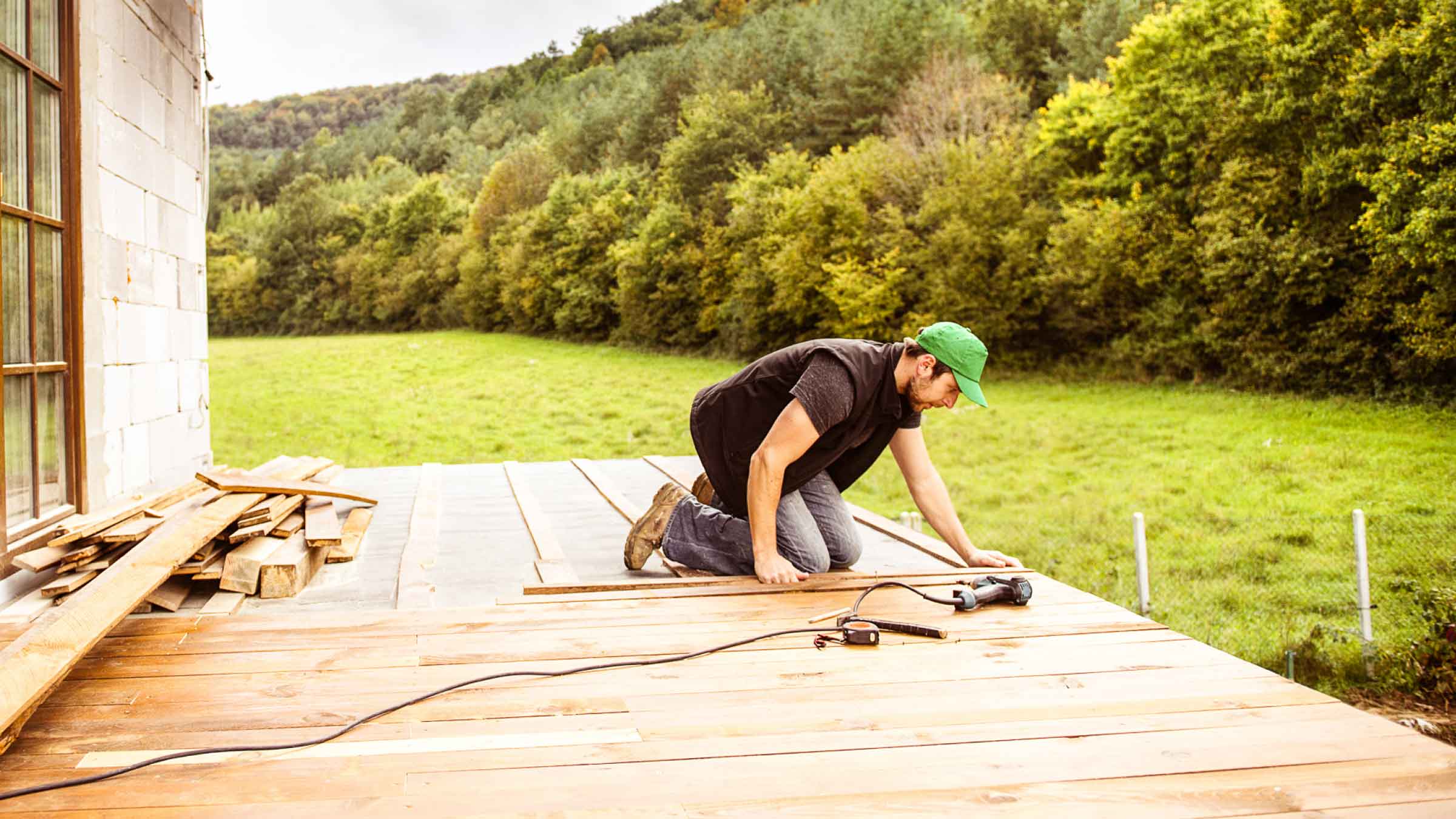 Tradie kneels on half finished decking, while measuring up some timber. It's out the back of a home, surrounded by green trees and a manicured lawn.