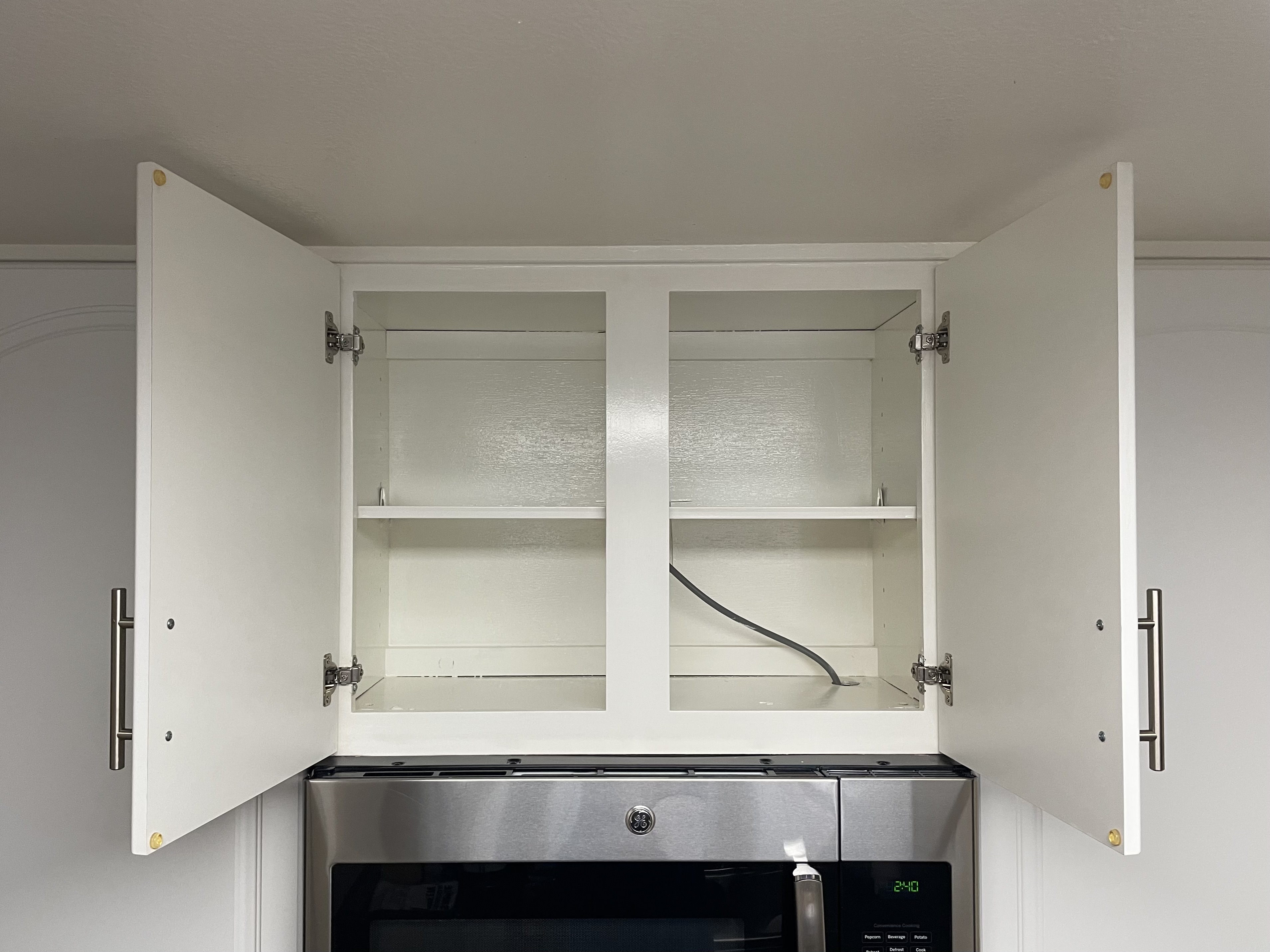 An open white kitchen cabinet above a microwave. The cabinet has two doors, both open to reveal empty shelves inside. There's a center divider and one visible shelf in each compartment. The cabinet doors have metal handles. A black power cord is visible on the bottom shelf. Below the cabinet, a stainless steel microwave with a digital display showing 2:40 can be seen.