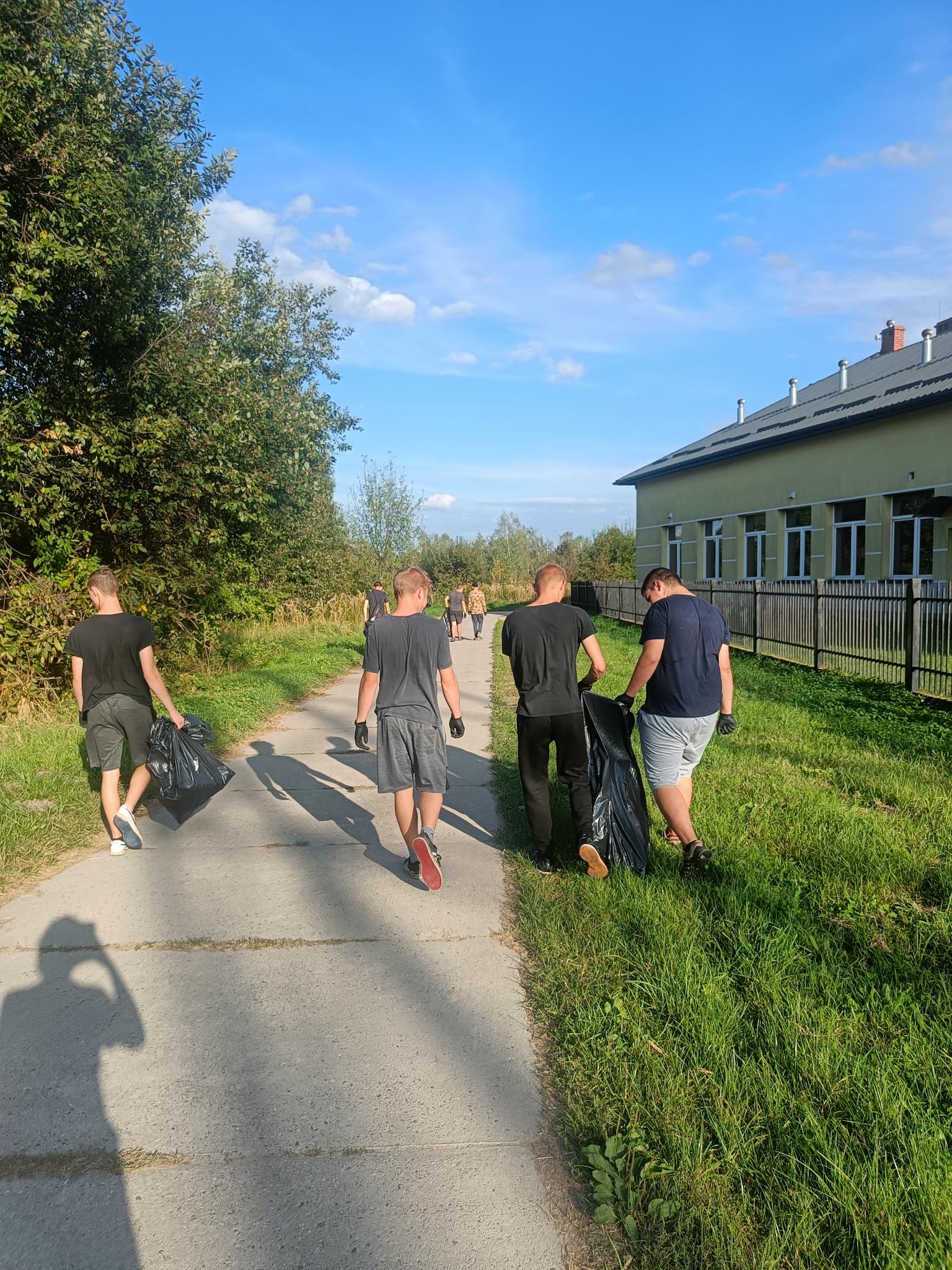 Sprzątanie świata | Wychowankowie MOW idą betonową drogą niosąc worki ze śmieciami, z lewej strony drzewa, z prawej trawa i żółty budynek z ogrodzeniem..jpg