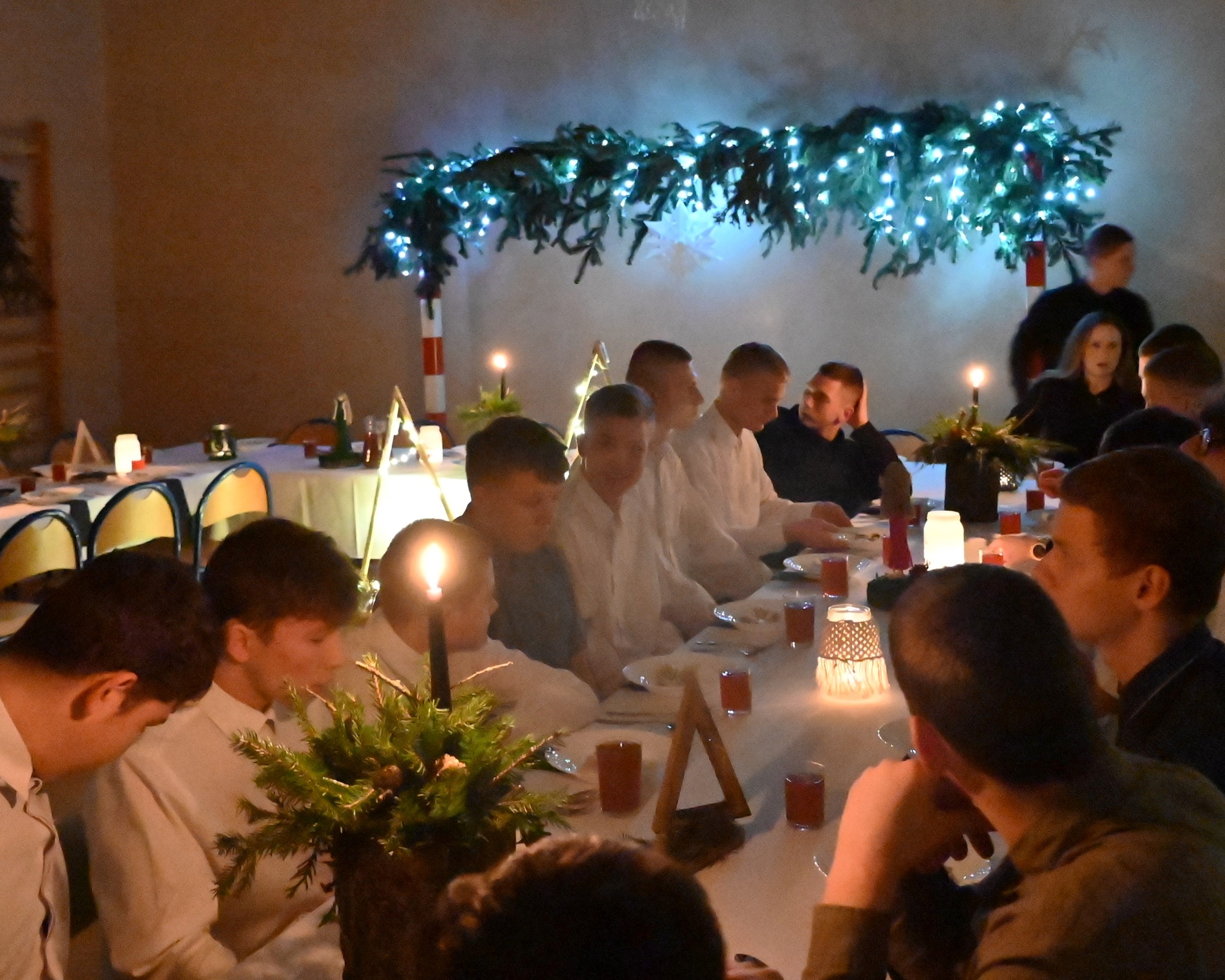 Wigilia w Ośrodku | Widok ogólny chłopców w białych koszulach zasiadających przy wigilijnym stole. W głębi stołu, między uczniami, jedna z wychowawczyń. Na stole i na ścianie stroiki ze śiatełkami bądź świecami.JPG