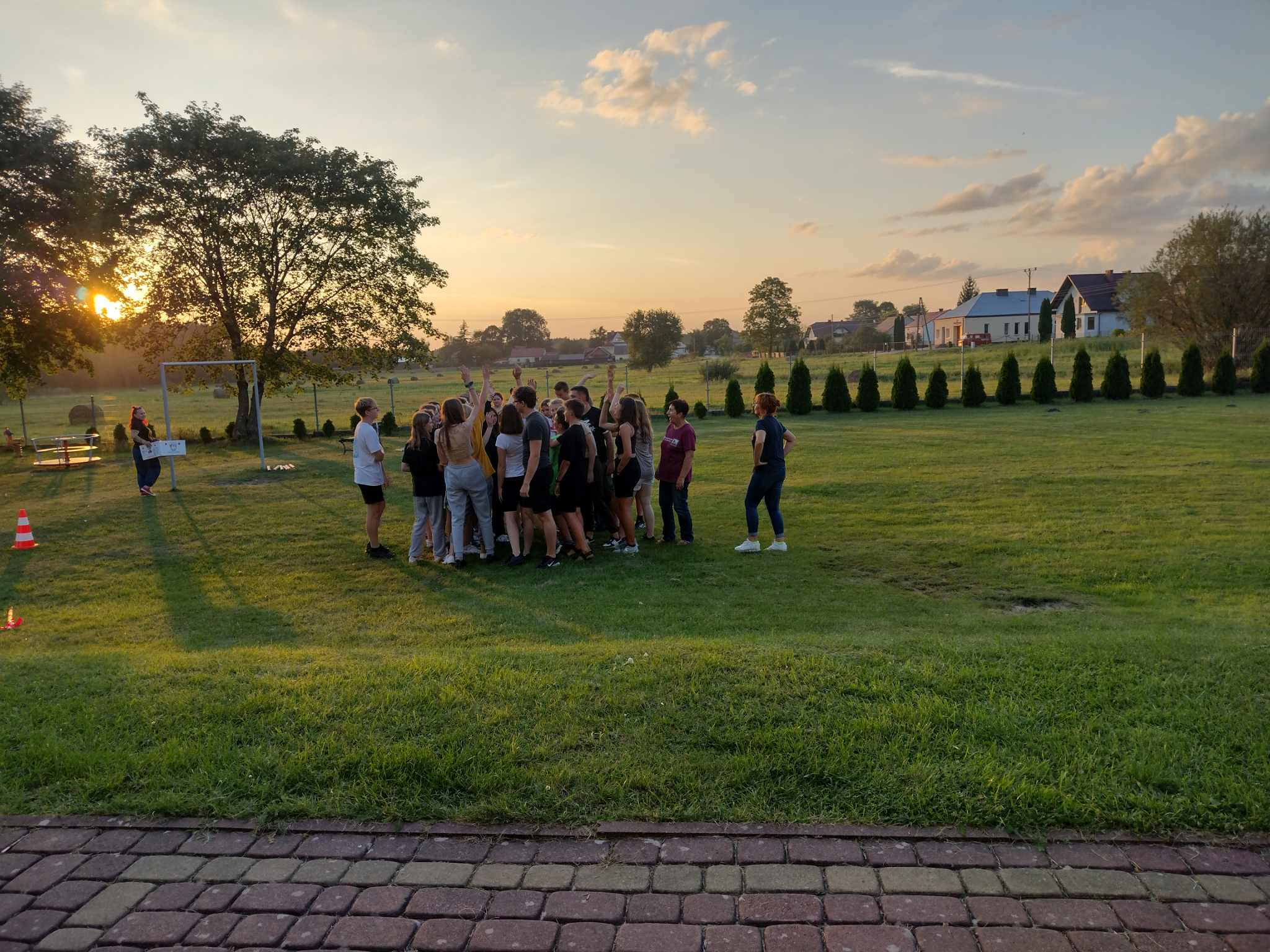Warsztaty profilaktyczne | Grupa młodzieży stoi blisko siebie na trawiastym boisku, część trzyma ręce podniesione w górę. Wszyscy są zwróceni twarzami do siebie. W tle zachodzące słońce..jpeg