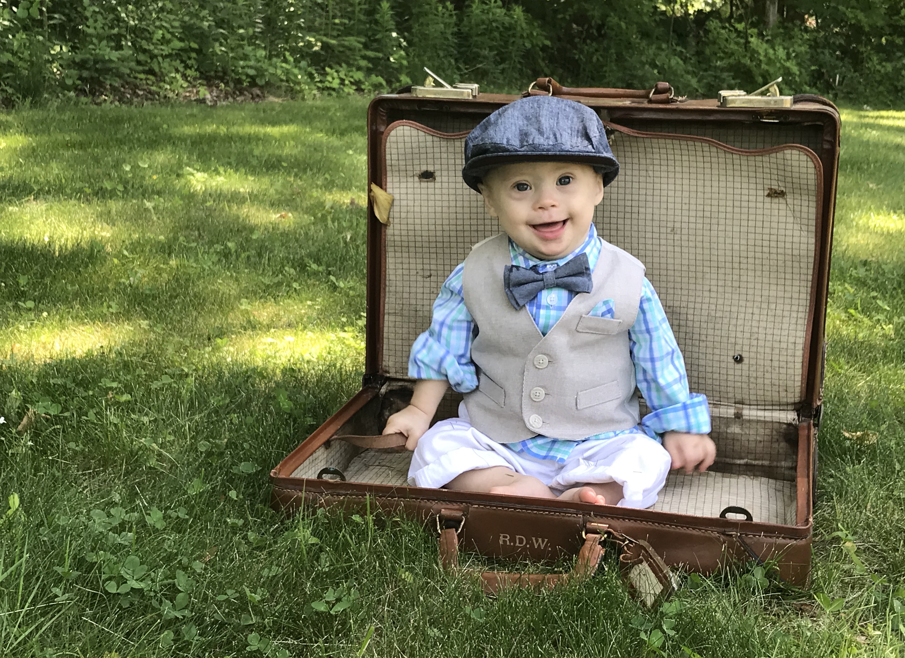 A baby with Down syndrome wearing a bowtie sitting in a briefcase in a field of grass