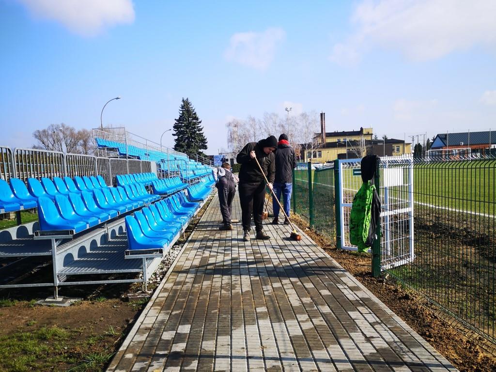 Porządki na stadionie | Trzy osoby ubrane w kurtki zamiatają chodnik z kostki brukowej. Po lewej niebieskie krzesełka trybun, z prawej ogrodzenie boiska.jpg