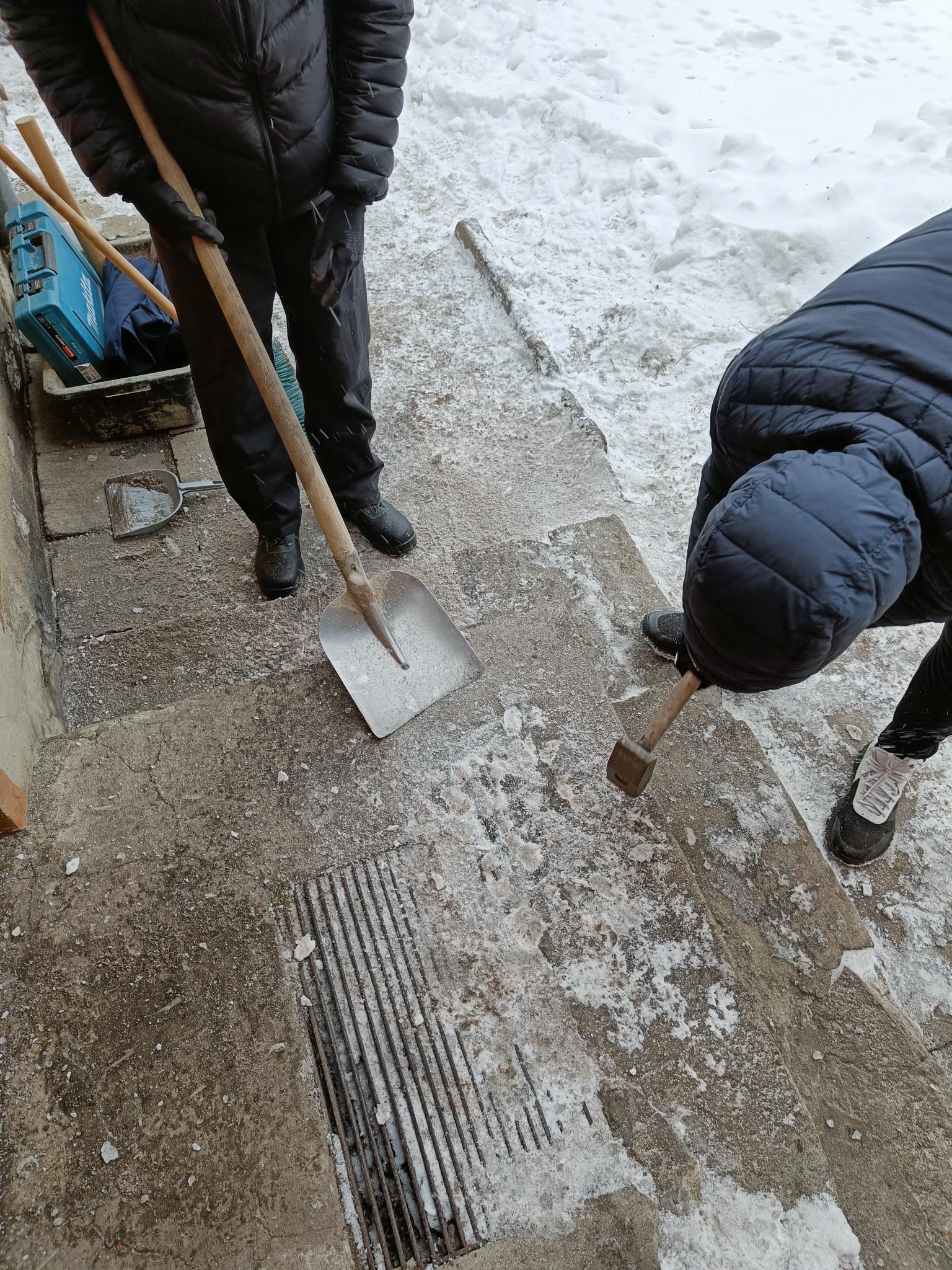 Wolontariat – pomoc w rozbiórce pieca | Dwóch chłopców skuwa lód na posadzce przed domem. Jeden rozbija skorupę młotkiem, drugi czeka z łopatą, by odgarnąć lód.jpg