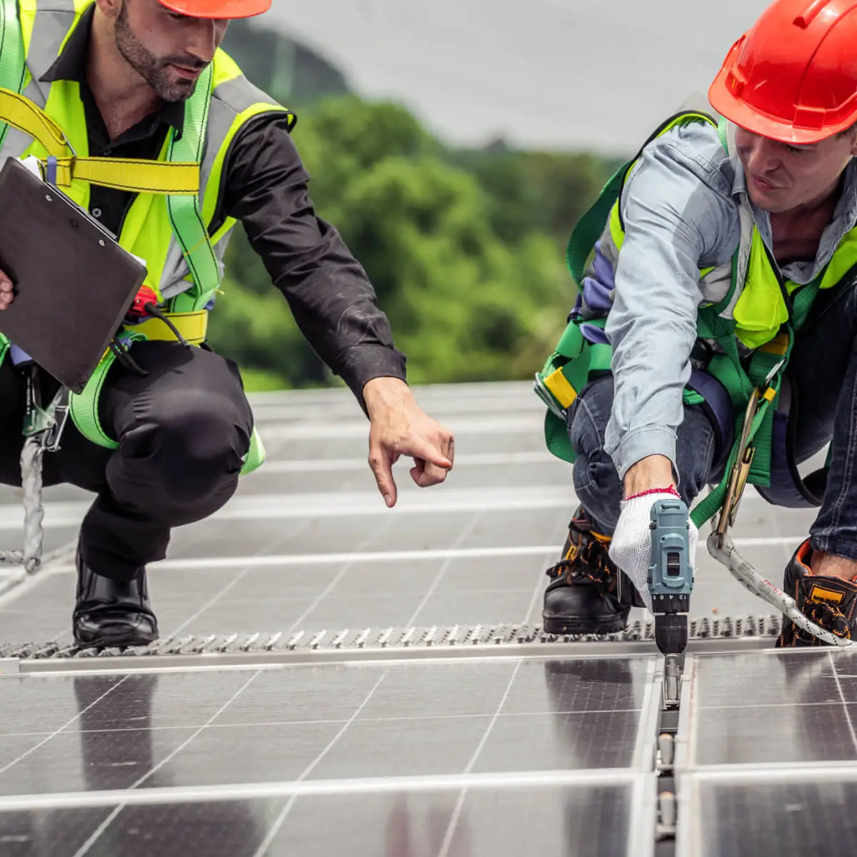 Two people stand on top of a solar panel covered roof. One person is pointing out fixes that need to be made from a checklist he has in his hand. The other person has a drill in one hand and is listening intently to the directions.
