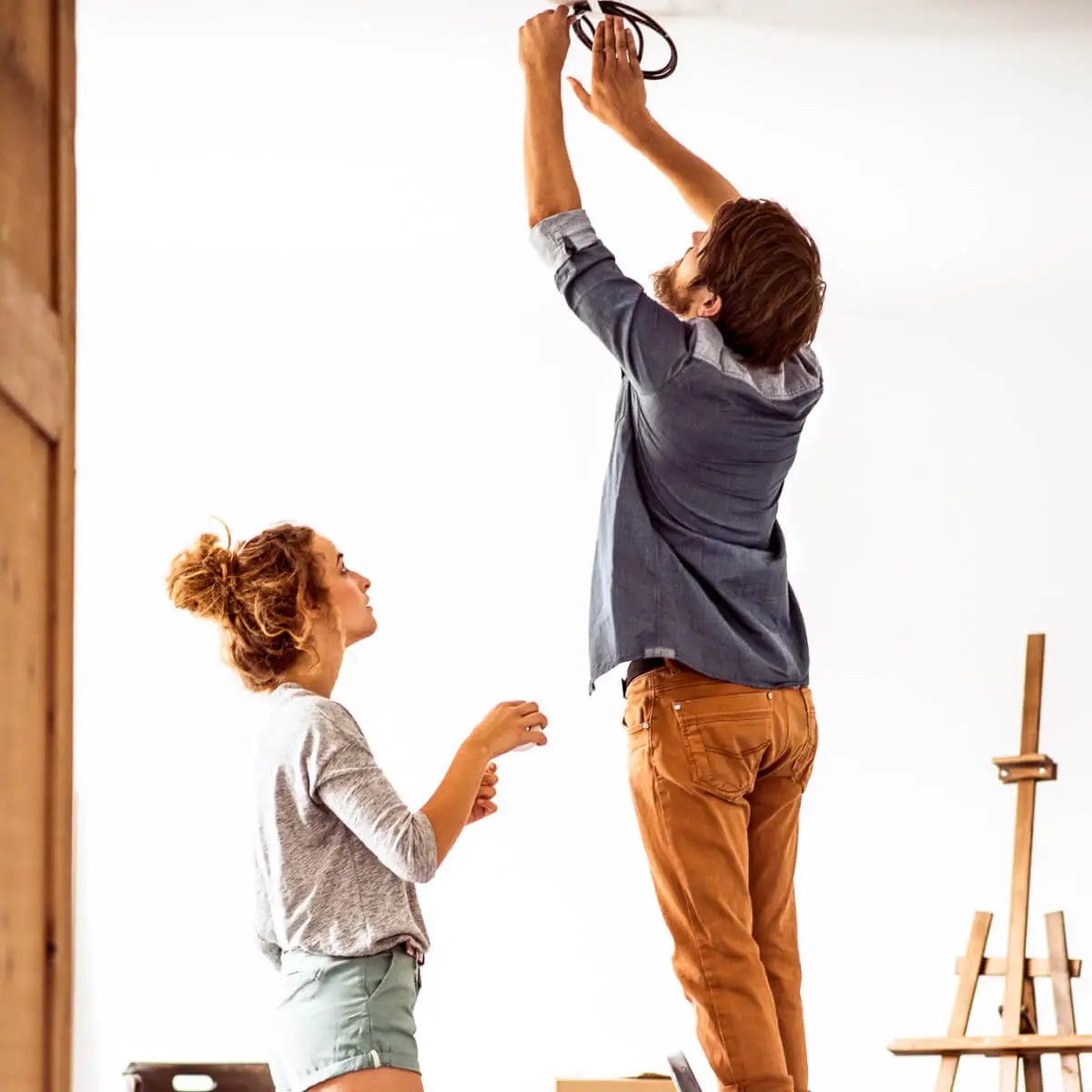 Young man stands on chair reaching up to electrical light wiring. A young woman stands beside him ready to help out.