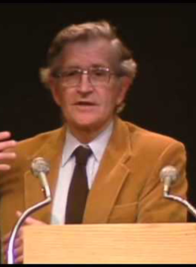 Noam Chomsky standing onstage behind a podium in front of a dark background