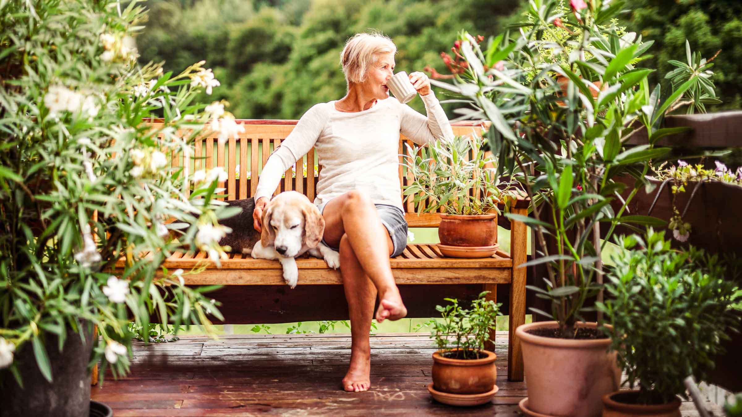 Woman sits on her outdoor bench surrounded by plants and trees, whilst sipping from a mug, and patting her sleepy dog by her side.