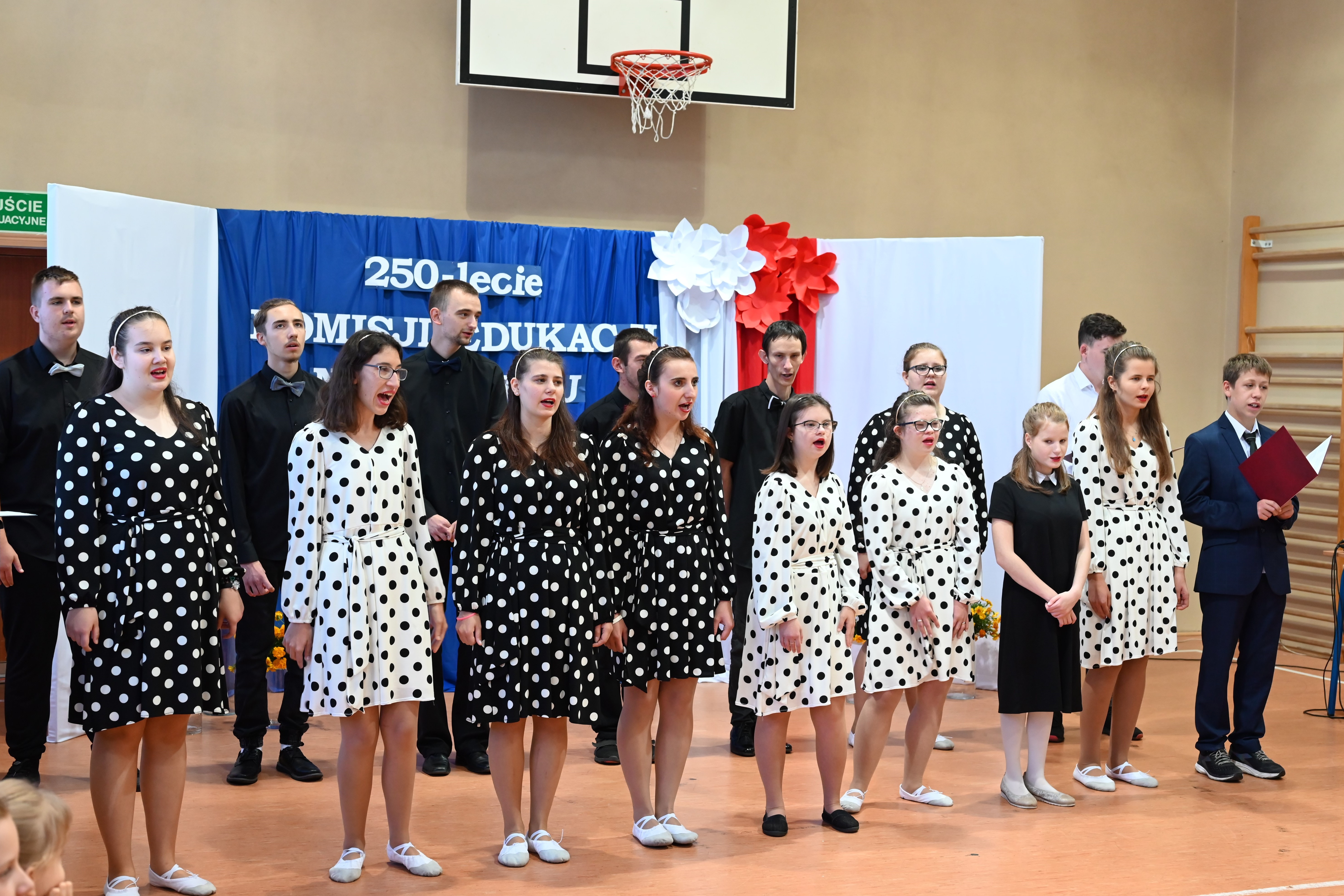 Dzień Edukacji Narodowej | Duża grupa młodzieży ubranej galowo chóralnie śpiewa piosenkę podczas alademii. Dziewczynki w białych sukienkach w czarne kropki lub w czarnych w białe kropki, chłopcy w czarnych lub białych koszulach, jeden w garniturze. W tle dekoracja..JPG