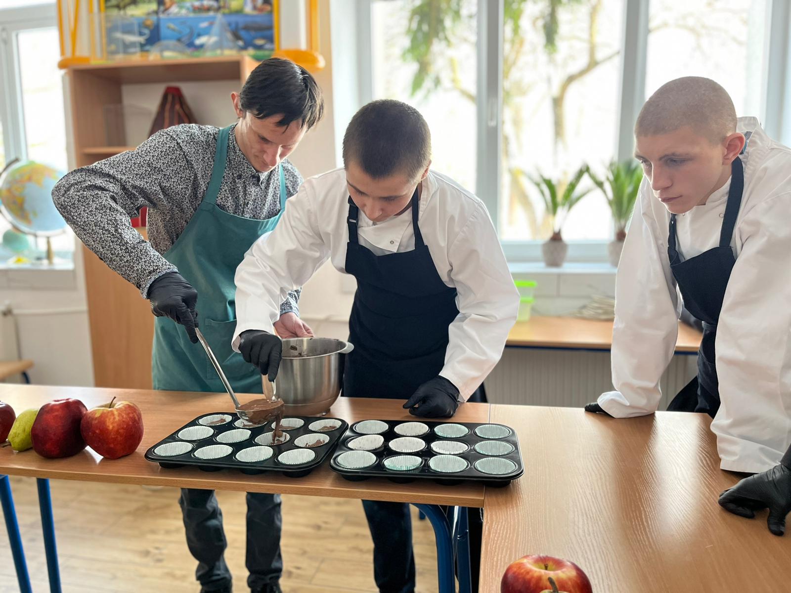 Przedświąteczne warsztaty kulinarne  | Dwóch chłopców nakłada chochlą i łyżeczką ciasto do foremek na babeczki. Obok stoi inny chłopiec przyglądając się pracy.jpg