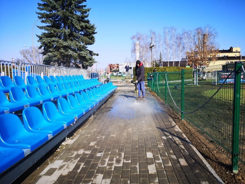 Porządki na stadionie | Chłopiec w kurtce z kapturem myje myjką ciśnieniową chodnik przy trybunach. Z lewej niebieskie krzesełka trybun, z prawej zielone ogrodzenie boiska.jpg