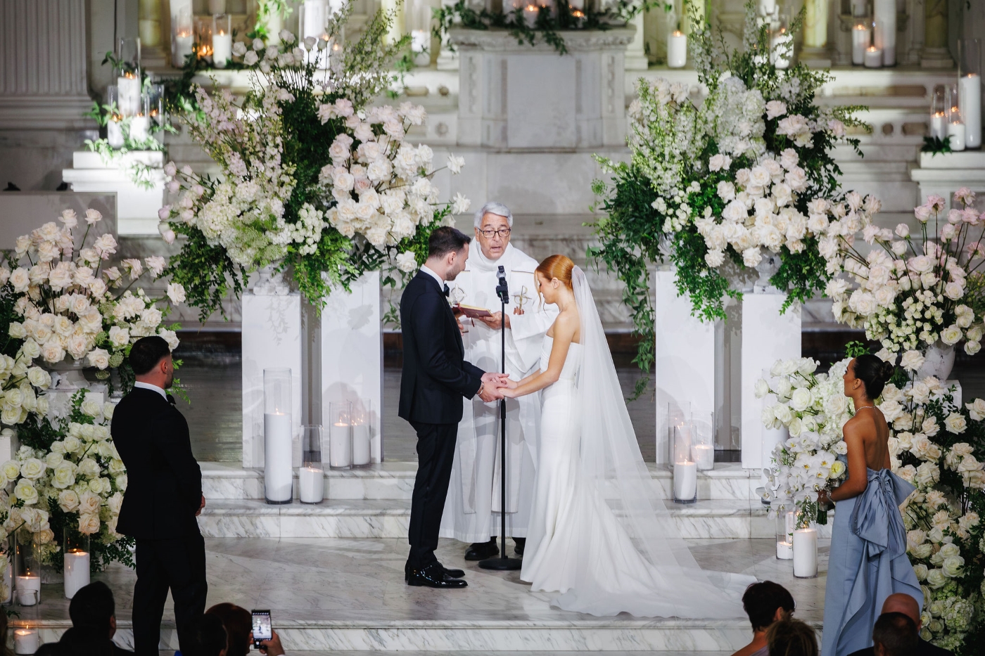 a bride and groom stand at the altar of their wedding ceremony, surrounded by white roses and their wedding guests and pastor