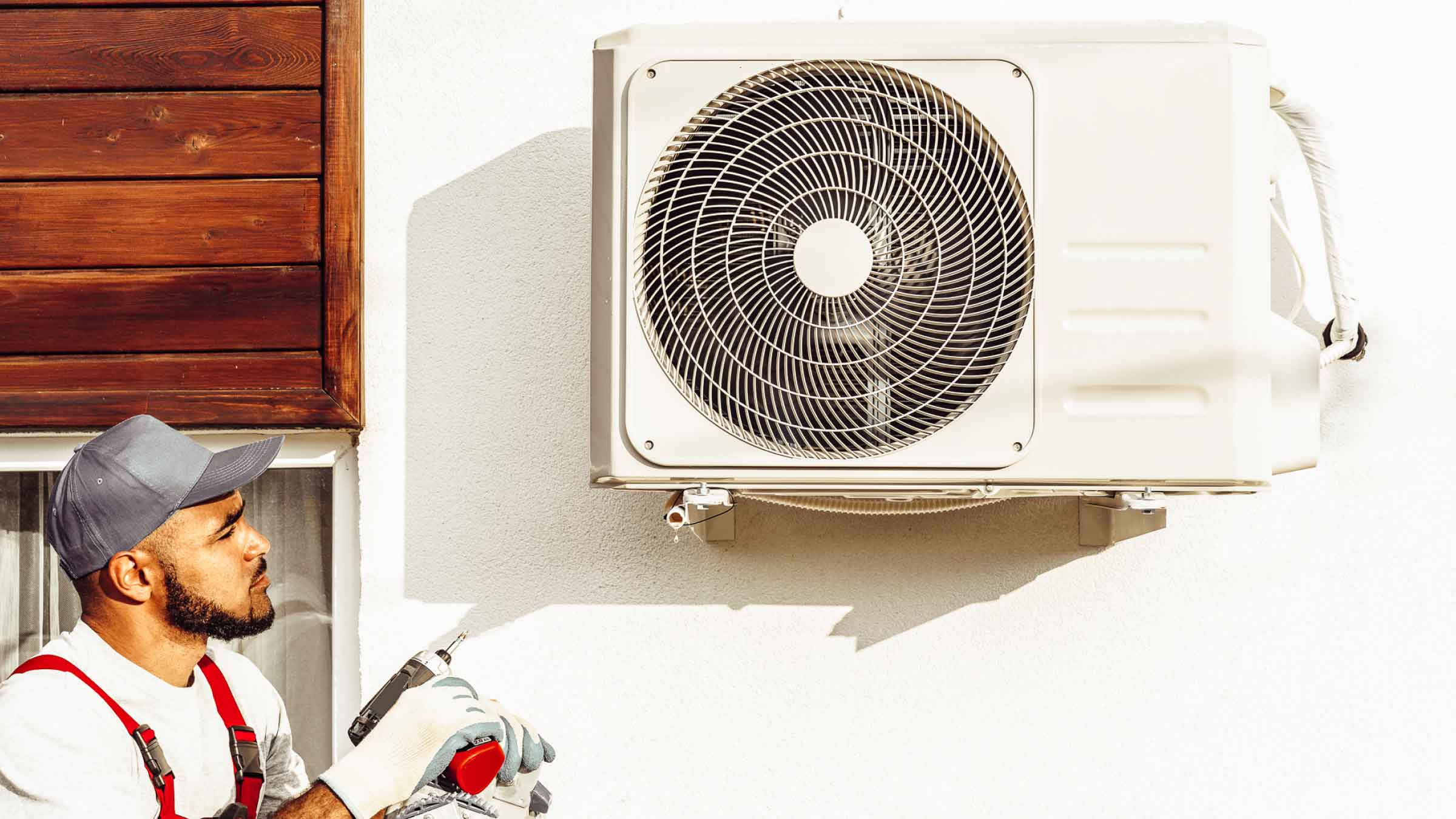 Air conditioner mounted to the outside wall of a home, man stands to the left with drill in hand.