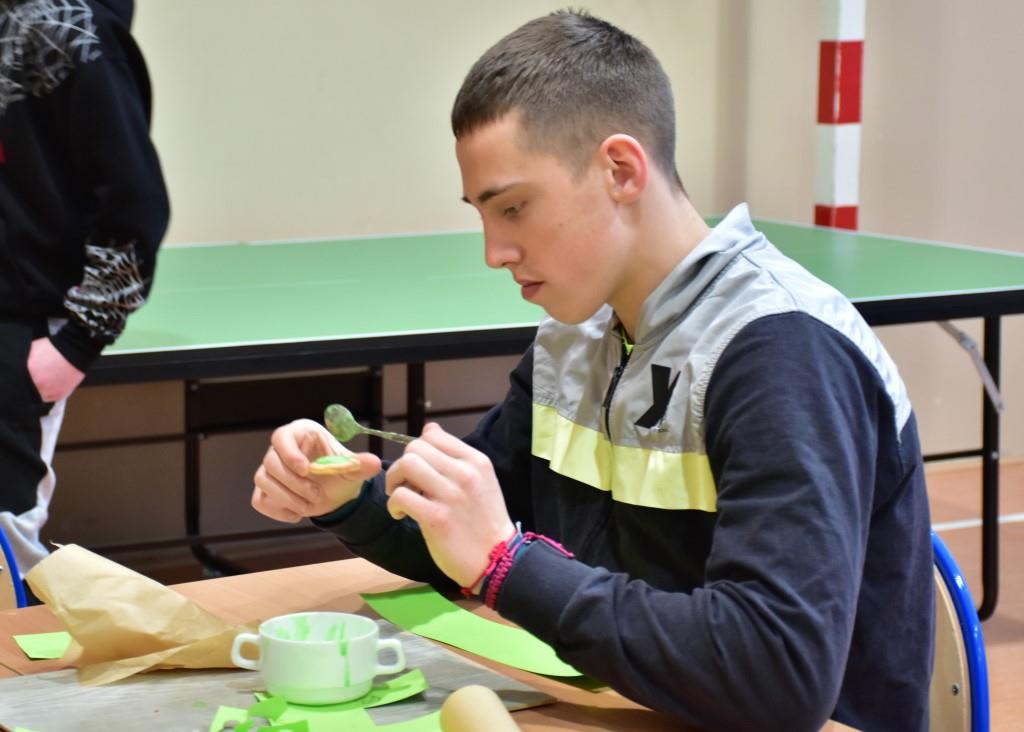 Dzień Św. Patryka  | Chłopiec siedzi przy stole, przed nim miseczka z zielonym lukrem. Chłopiec przy pomocy łyżeczki ozdabia lukrem ciasteczko.JPG