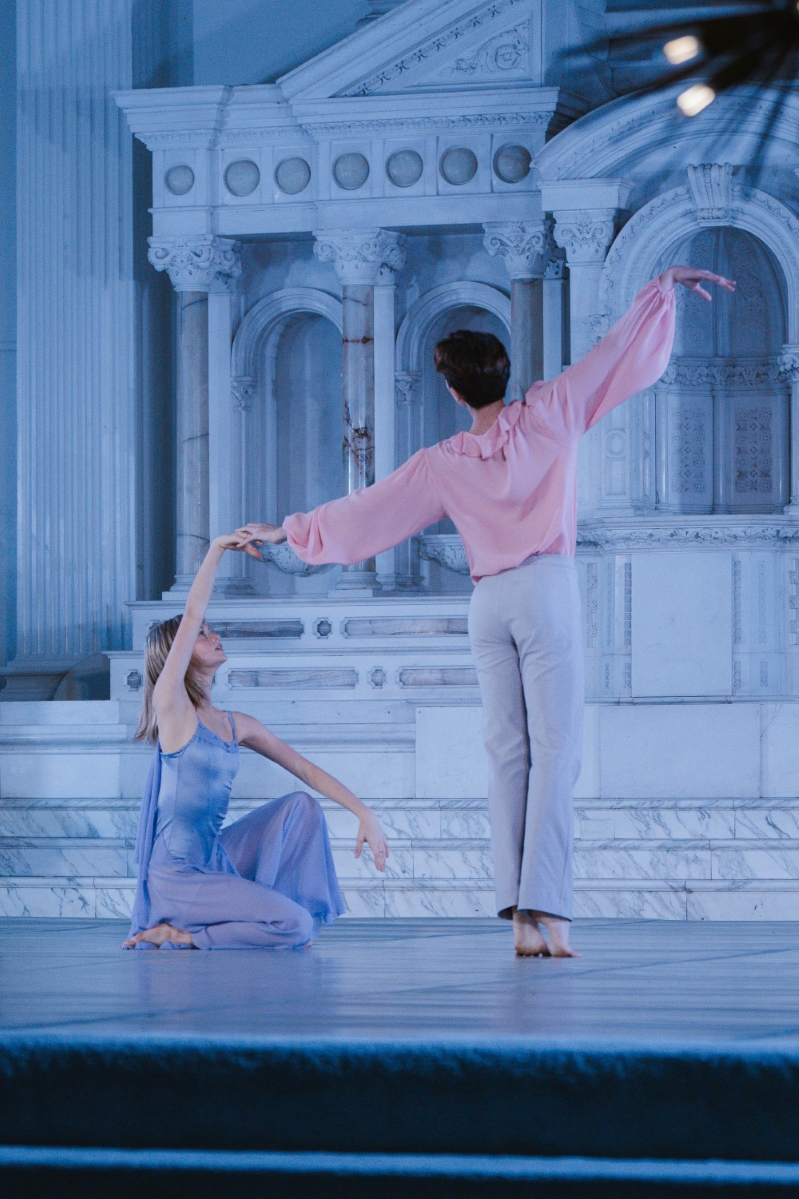 a female dancer dressed in blue sits on the ground, holding the hand of a male dancer in pink, at a performance at a gala at Vibiana in Downtown Los Angeles