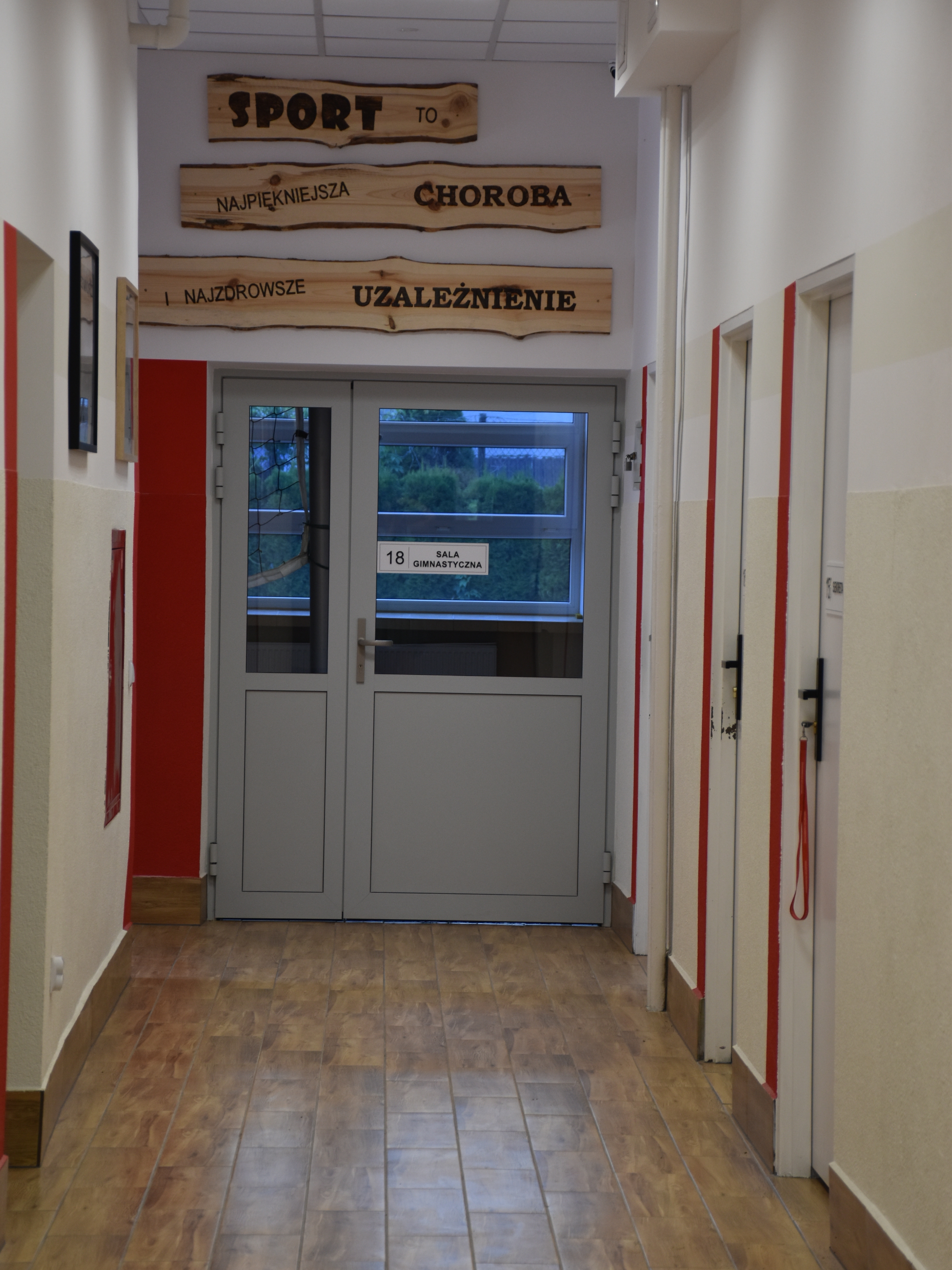 Uroczyste podsumowanie projektu „Dostępna Szkoła” | Korytarz szkolny w MOW prowadzący na salę gimnastyczną, w głębi korytarza drzwi od sali oznakowane czerwonym pasem, na drzwiami napis wypalony na desce..JPG