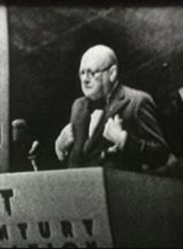 Grainy black and white image of Winston Churchill on a stage walking to podium as two men onstage