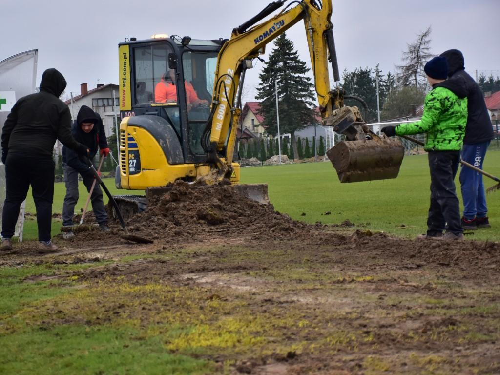 Porządki na stadionie | Żółta, nieduża koparka stoi na murawie boiska, obok niej cztery osoby z grabiami i łopatami w rękach.JPG