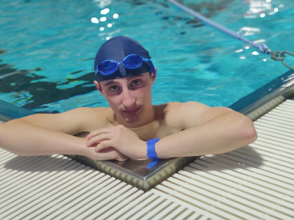 Zajęcia czasie wolnym | Wychowanek MOW w niebieskim czepku i okularach do pływania na czole podczas kąpieli w basenie. Chłopiec opiera się rękami o brzeg basenu i patrzy prosto w obiektyw aparatu..jpg