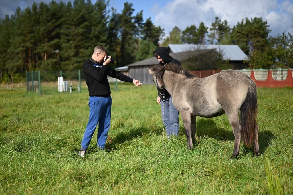 Obóz w Hucie Różanieckiej | Dwóch wychowanków MOW stoi na łące obok konika, jeden z nich robi mu zdjęcie wyciągając jednocześnie do niego rękę. Z tyłu widoczne ogrodzenie, za nim budynek, w tle drzewa..JPG