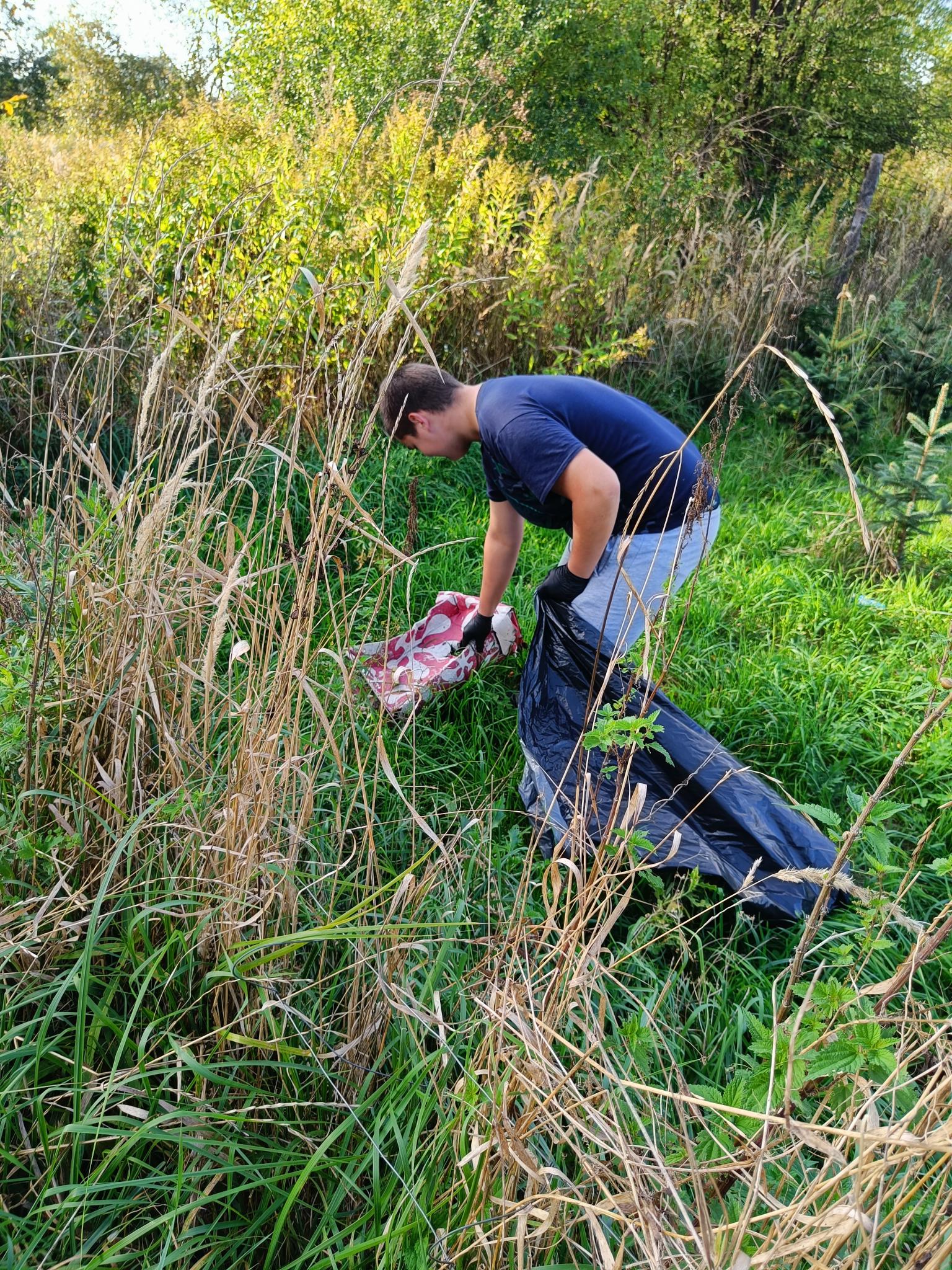 Sprzątanie świata | Wychowanek MOW pochyla się między wysokimi trawami podnosząc śmieci. W rece trzyma duży czarny worek na odpady..jpg