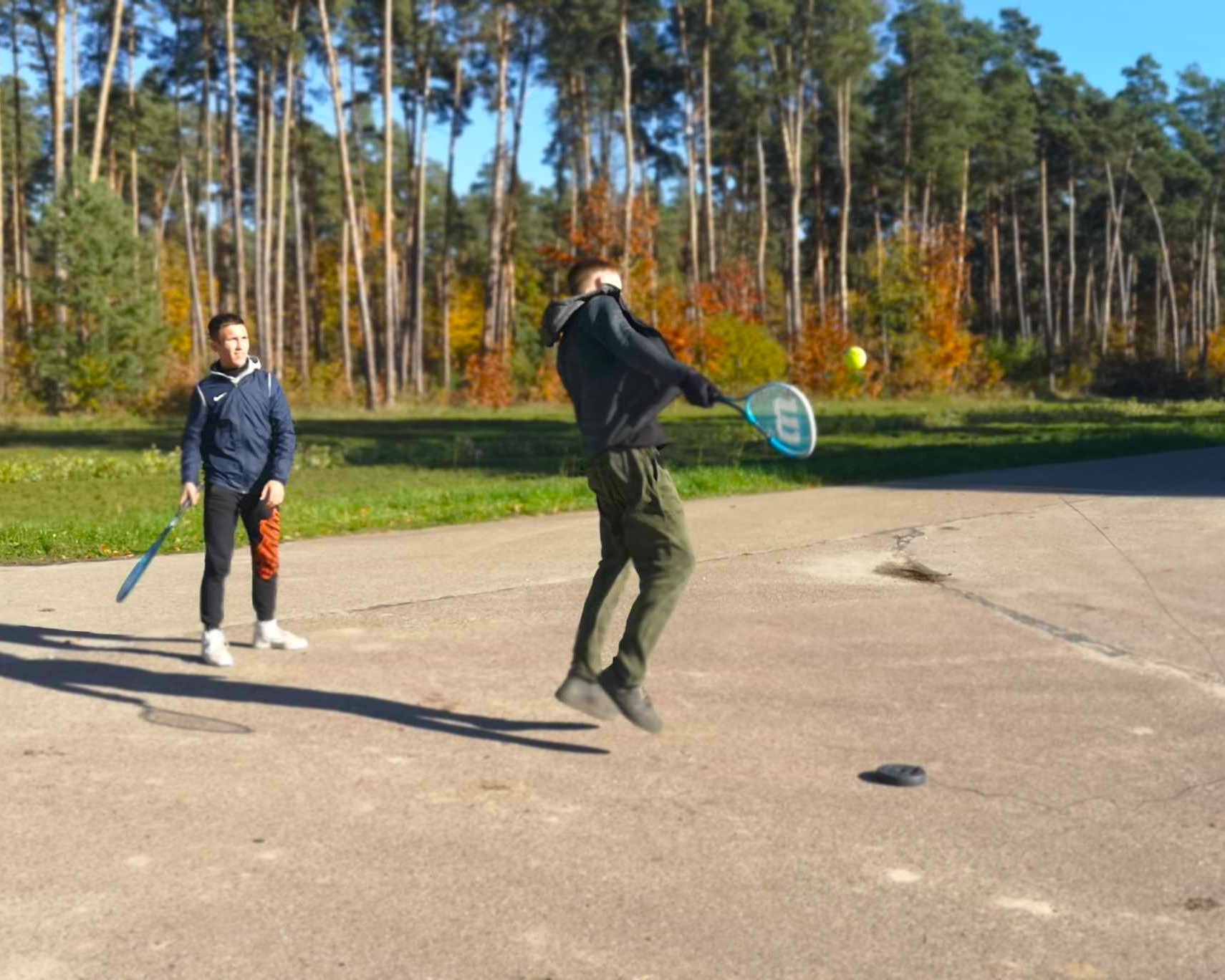 Zajęcia w szkółce leśnej | Dwóch chłopców z rakietkami, podczas gry na placu. Jeden jest w trakcie wyskoku, tuż przed odbiciem piłki w locie. W tle jesienny las..jpg