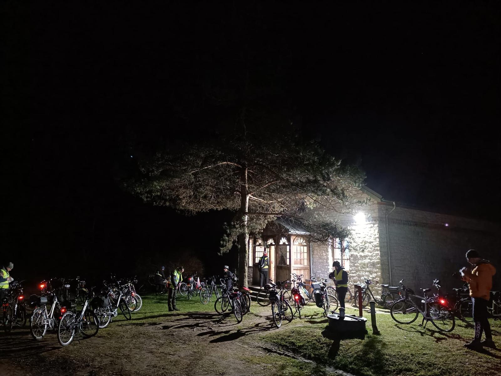 Rowerowa Droga Krzyżowa | Duża, murowana kaplica oświetlona latarnią. Przed nią drzewo i kilkanaście zaparkowanych rowerów, między nimi stoi kilka osób.Zdjęcie nocne.jpg