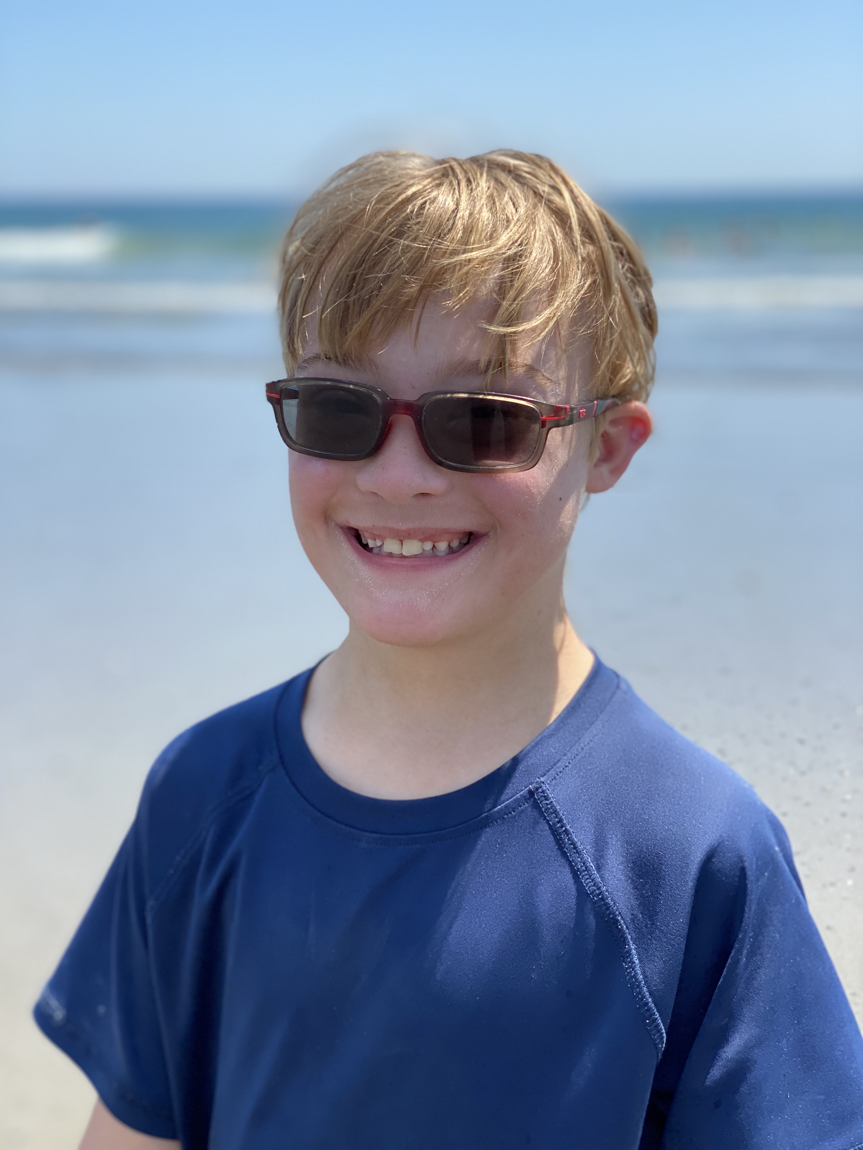 A young boy with down syndrome smiling at the beach wearing sunglasses