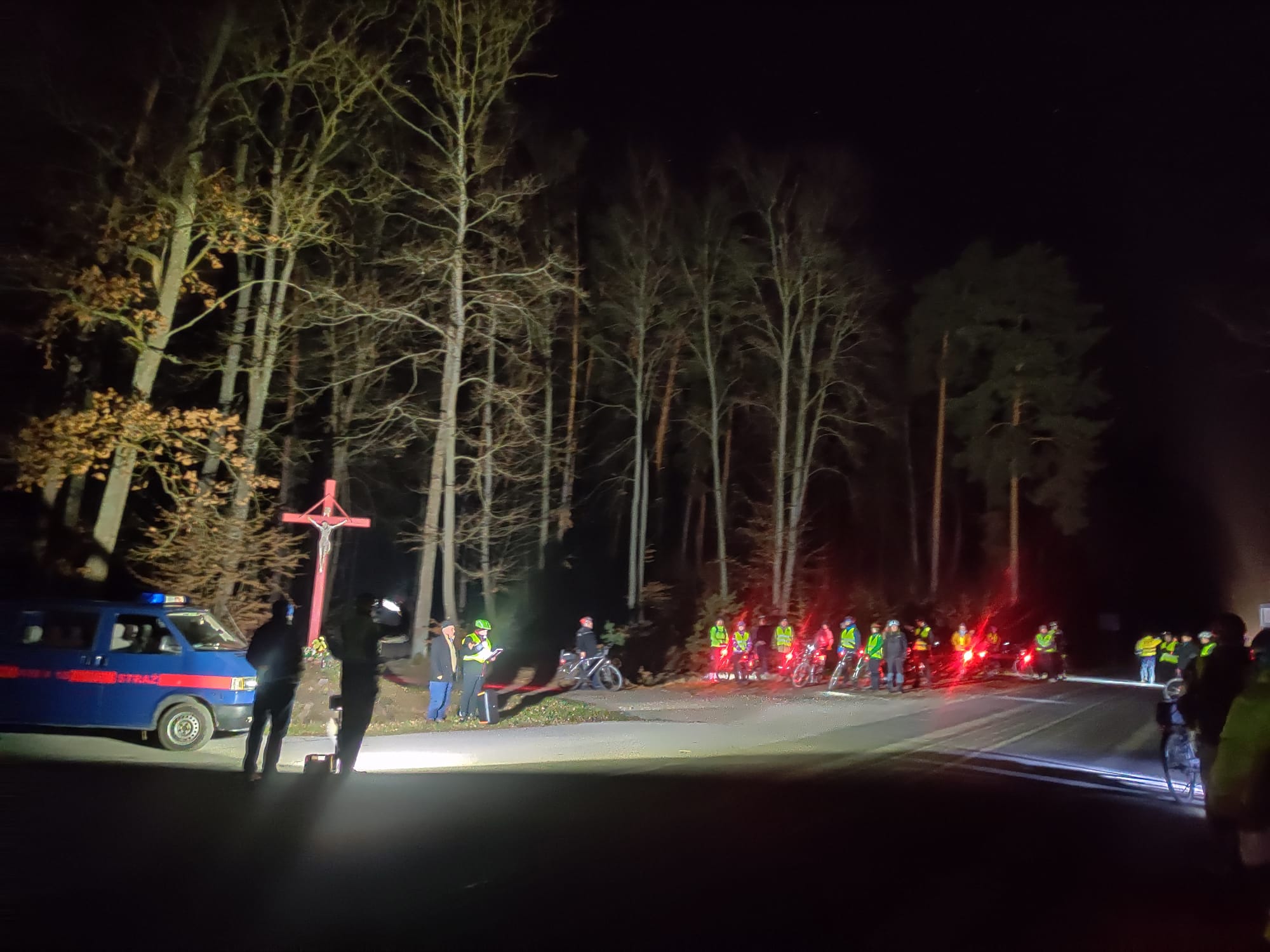 Rowerowa Droga Krzyżowa | Las nocą, drzewa oświetlone reflektorami. Między drzewami duży czerwony krzyż. Obok grupa kolarzy ze swoimi rowerami, z boku samochód straży.jpg