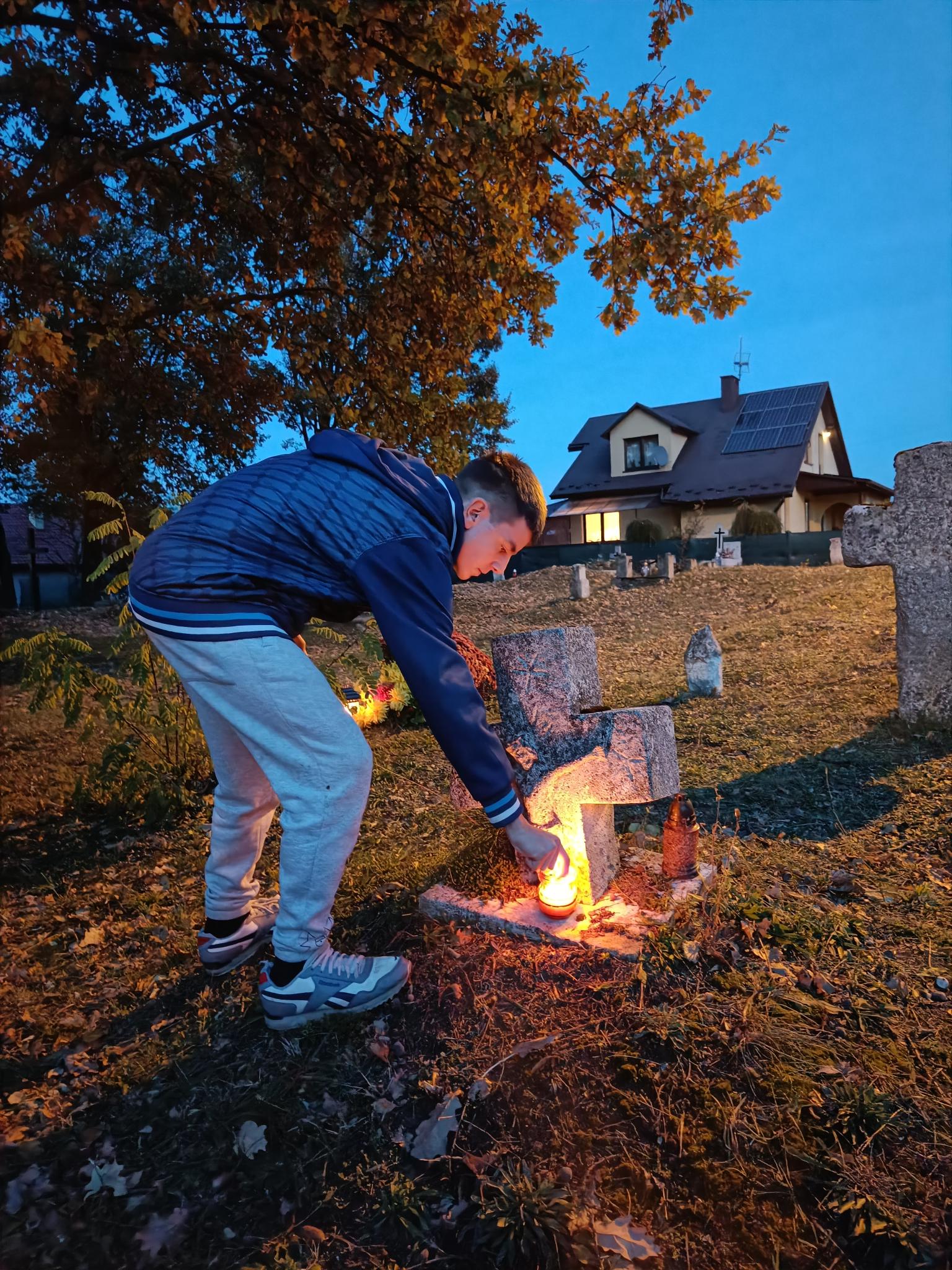 Pamiętamy... | Chłopiec pochyla się stawiając na ziemi pod kamiennym, niskim krzyżem zapalony znicz. W tle  inne krzyże i nagrobki, galąź liściastego drzewa pochylająca się do ziemi. W oddali dom z zapalonym w oknie światłem. Zdjęcie robione o świcie..jpg