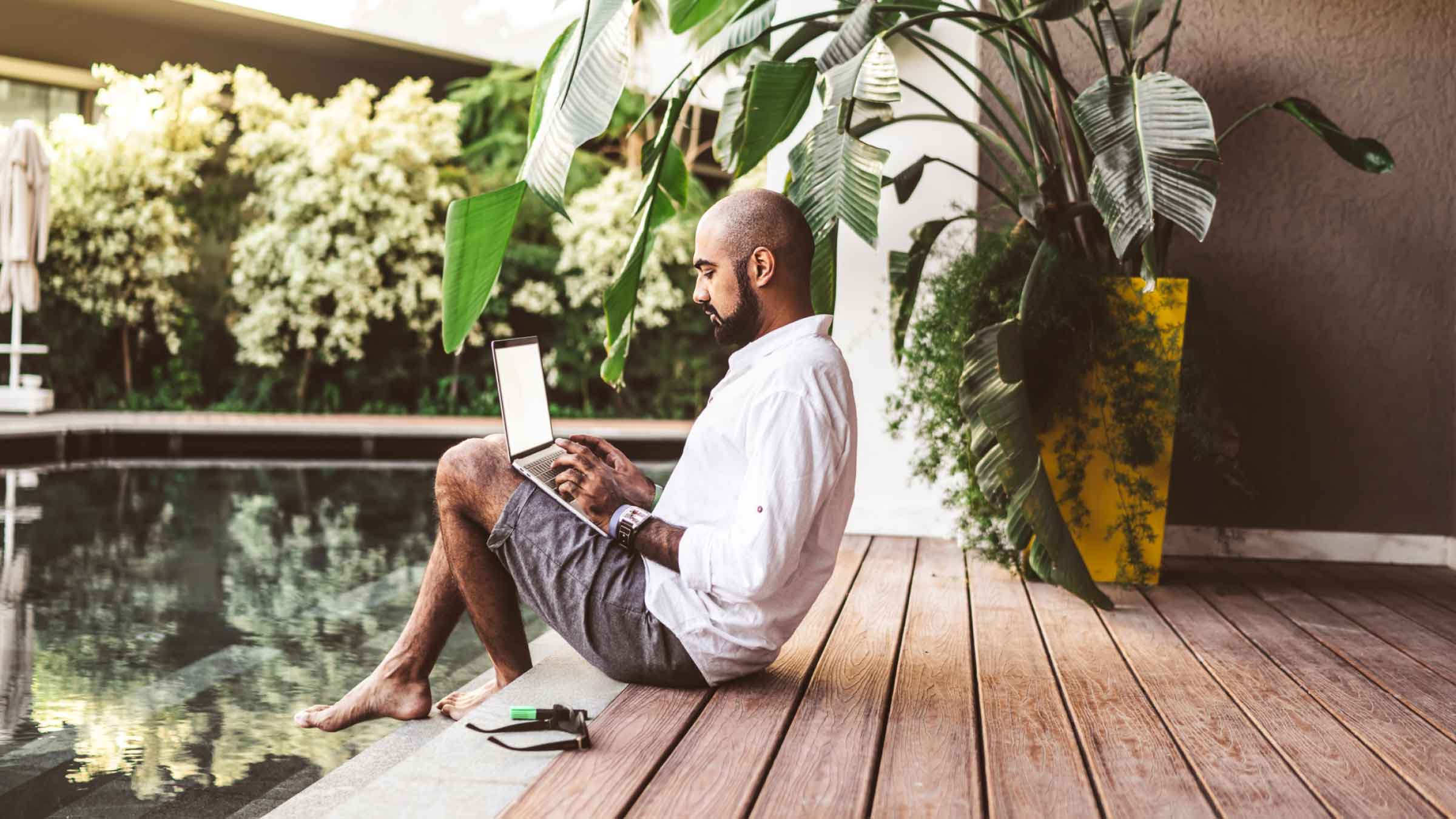 Man sits on the edge of a pool surrounded by wooden decking, palm leaves dangle in the background. He's about to dip his toe in the water while he works on his laptop.