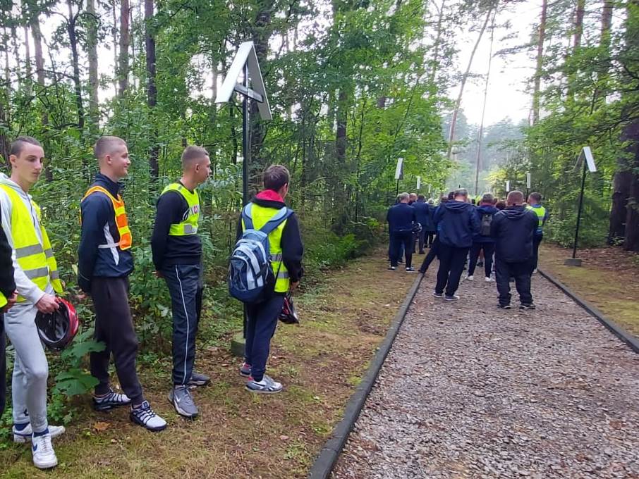 Rajd rowerowy | Wychowankowie MOW stoją przy leśnej ścieżce, w głębi po ścieżce idą ludzie, z obu stron drzewa i krzewy..jpg