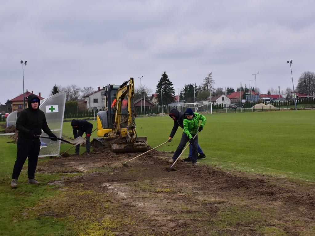 Porządki na stadionie | Trzy osoby grabią ziemię, jedna patrzy w stronę obiektywu. Między nimi zółta koparka.JPG