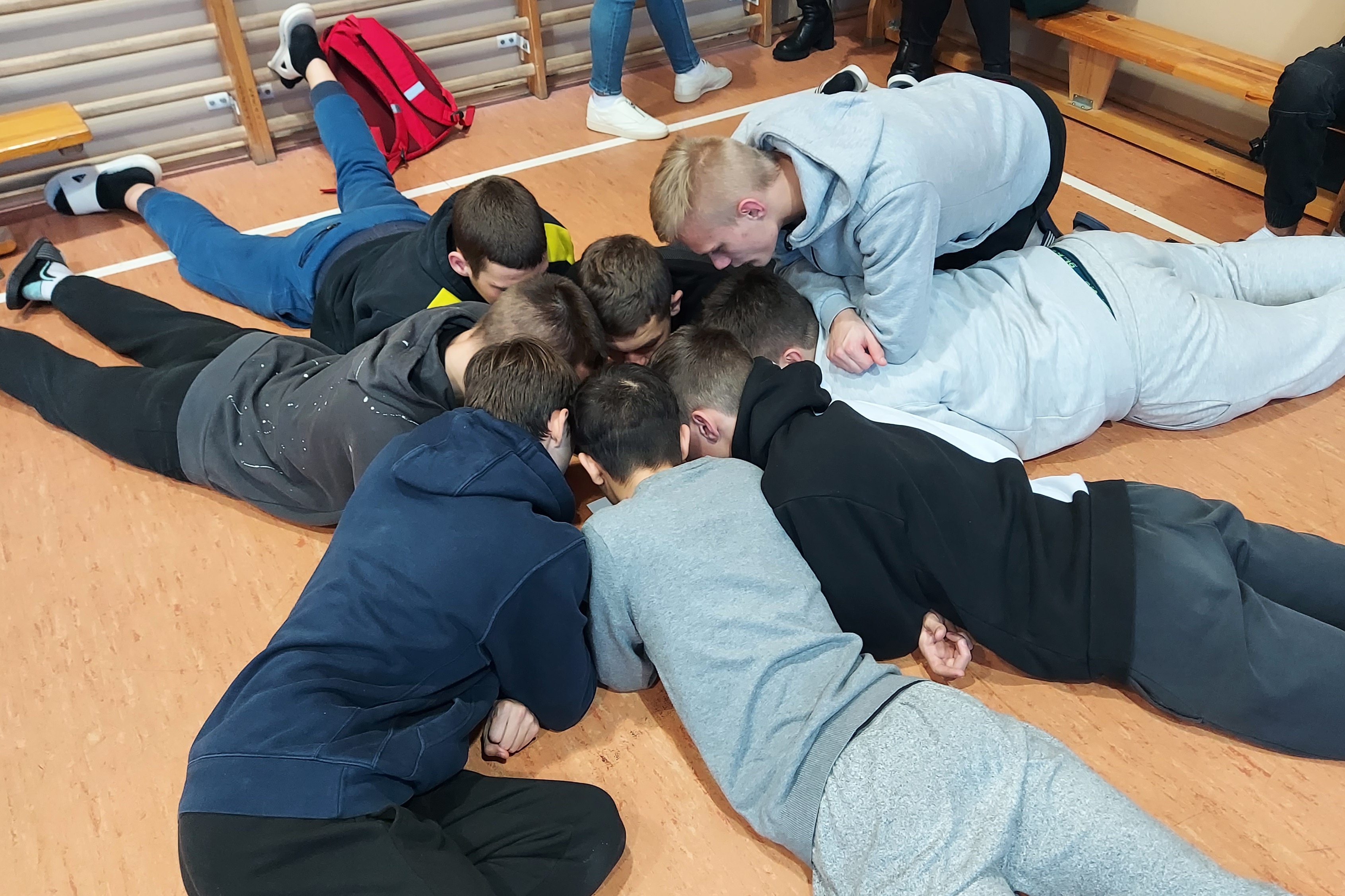 Turniej Wiedzy i Sprawności – edycja Mikołajkowa. | Chłopcy leżą na podłodze na burzuchach, głowy bardzo blisko siebie. Między nimi kartka z zadaniem.jpg