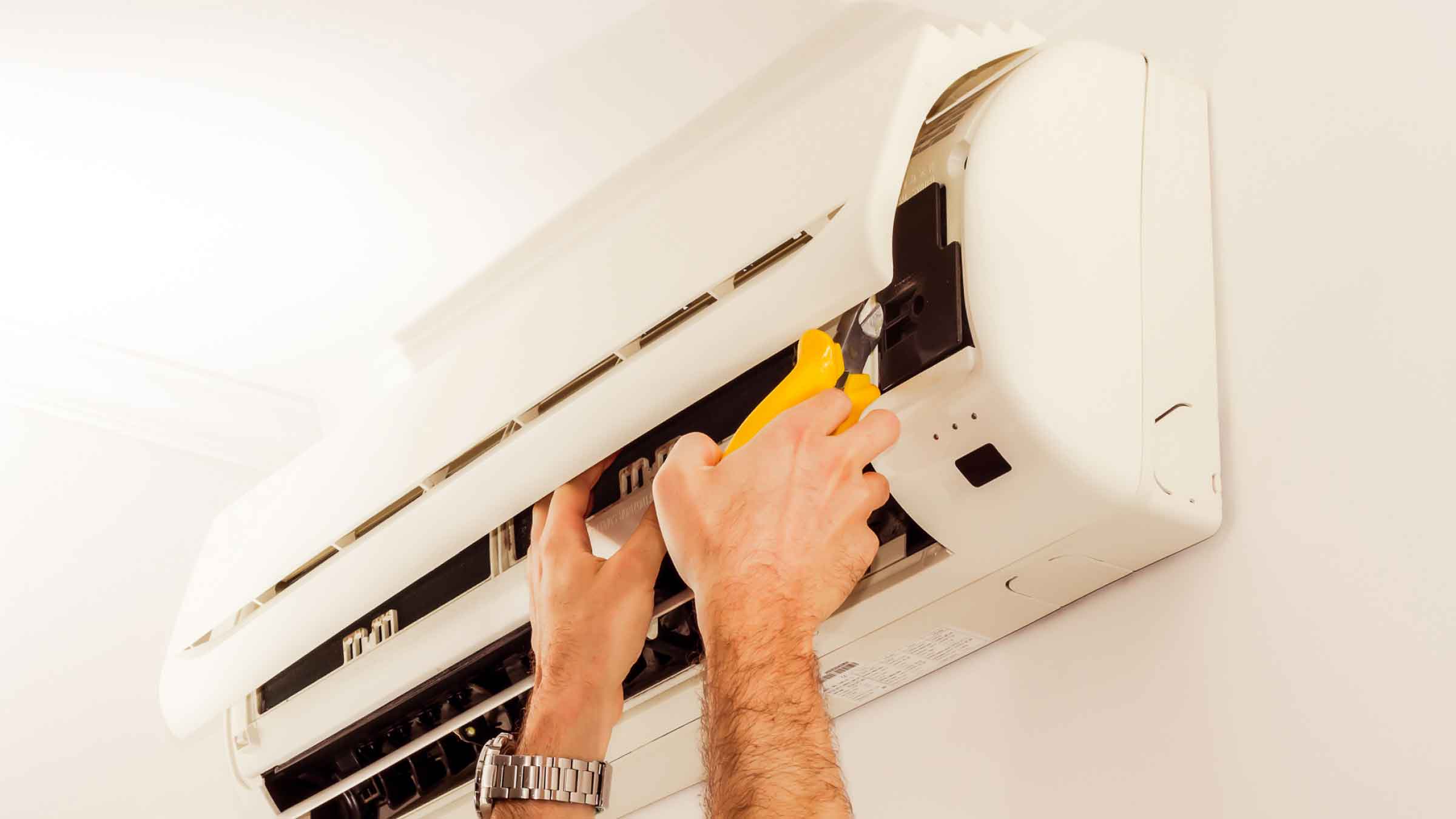 And air conditioner mounted on indoor wall, with two hands trying to fix it, one holding a yellow set of pliers.