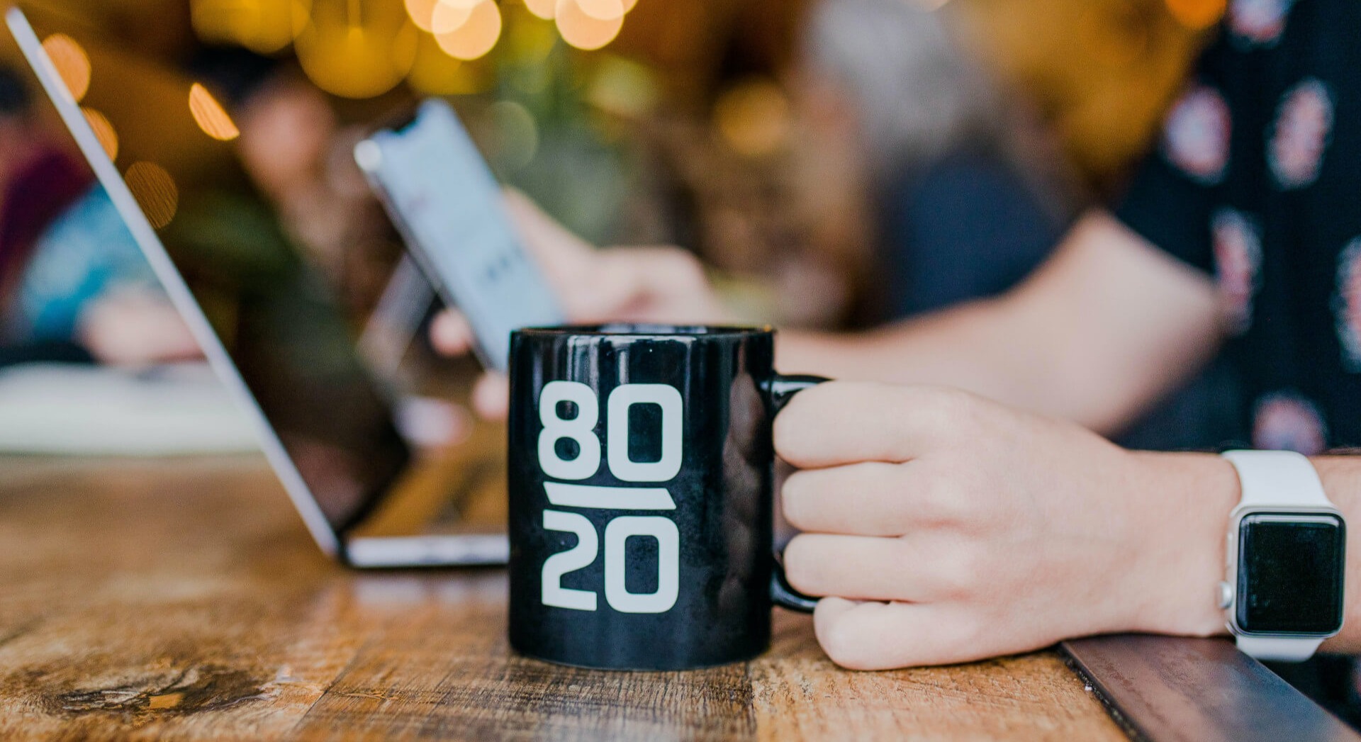 Blick von der Seite auf einen Tisch auf dem eine Tasse mit der Aufschrift "80/20" steht und gerade von einer Person mit Apple Watch am Handgelenk gegriffen wird. Im Hintergrund steht ein aufgeklappter Laptop. Mit automatisierter Buchhaltung kannst du nicht nur Zeit sondern auch Geld sparen. Wir zeigen dir wie!