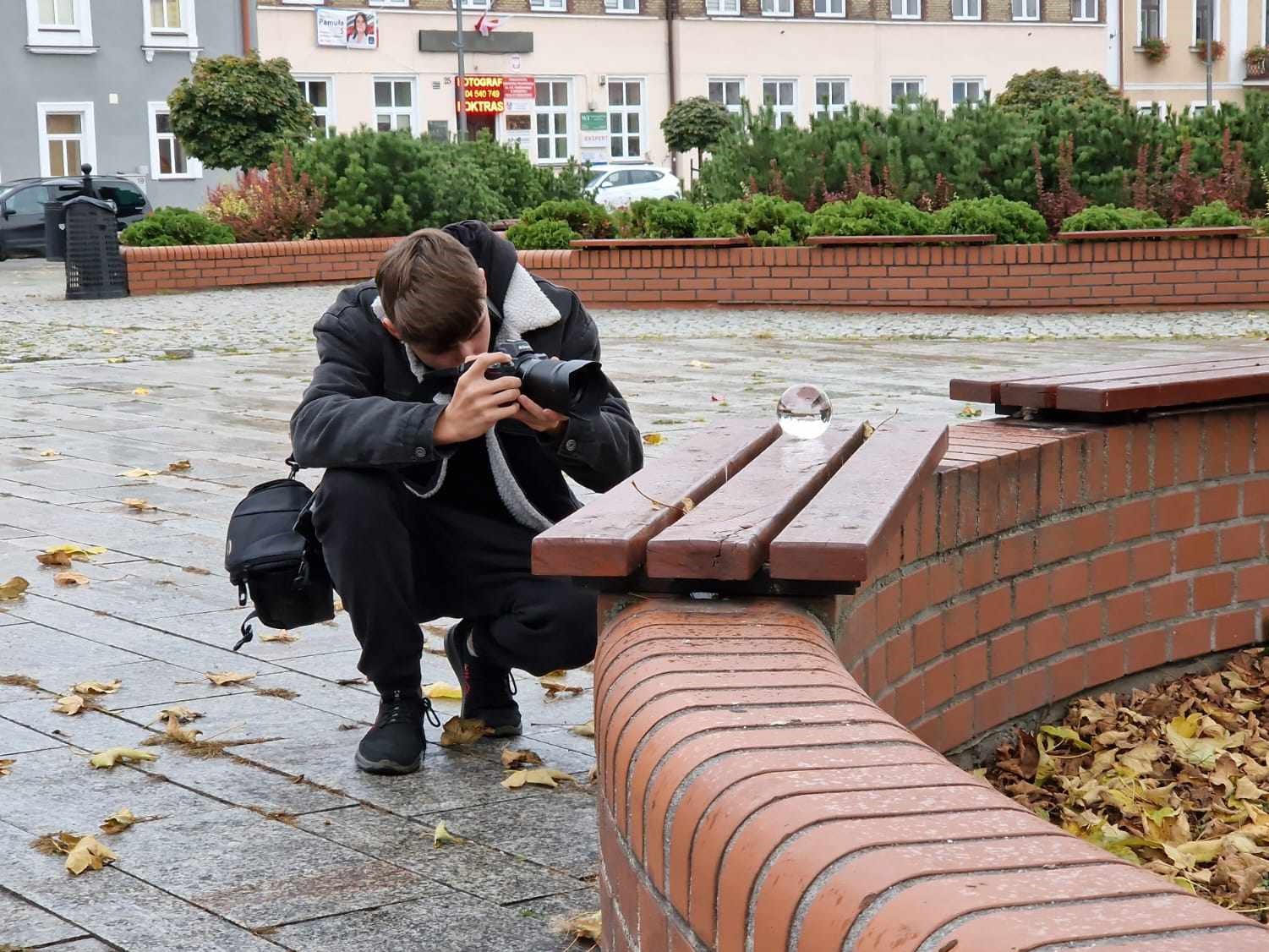 Fotografia jeszcze raz | Chłopiec z aparatem kuca fotografując szklaną kulę leżącą na deskach ławki. W tle krzewy i zabudowania..jpg