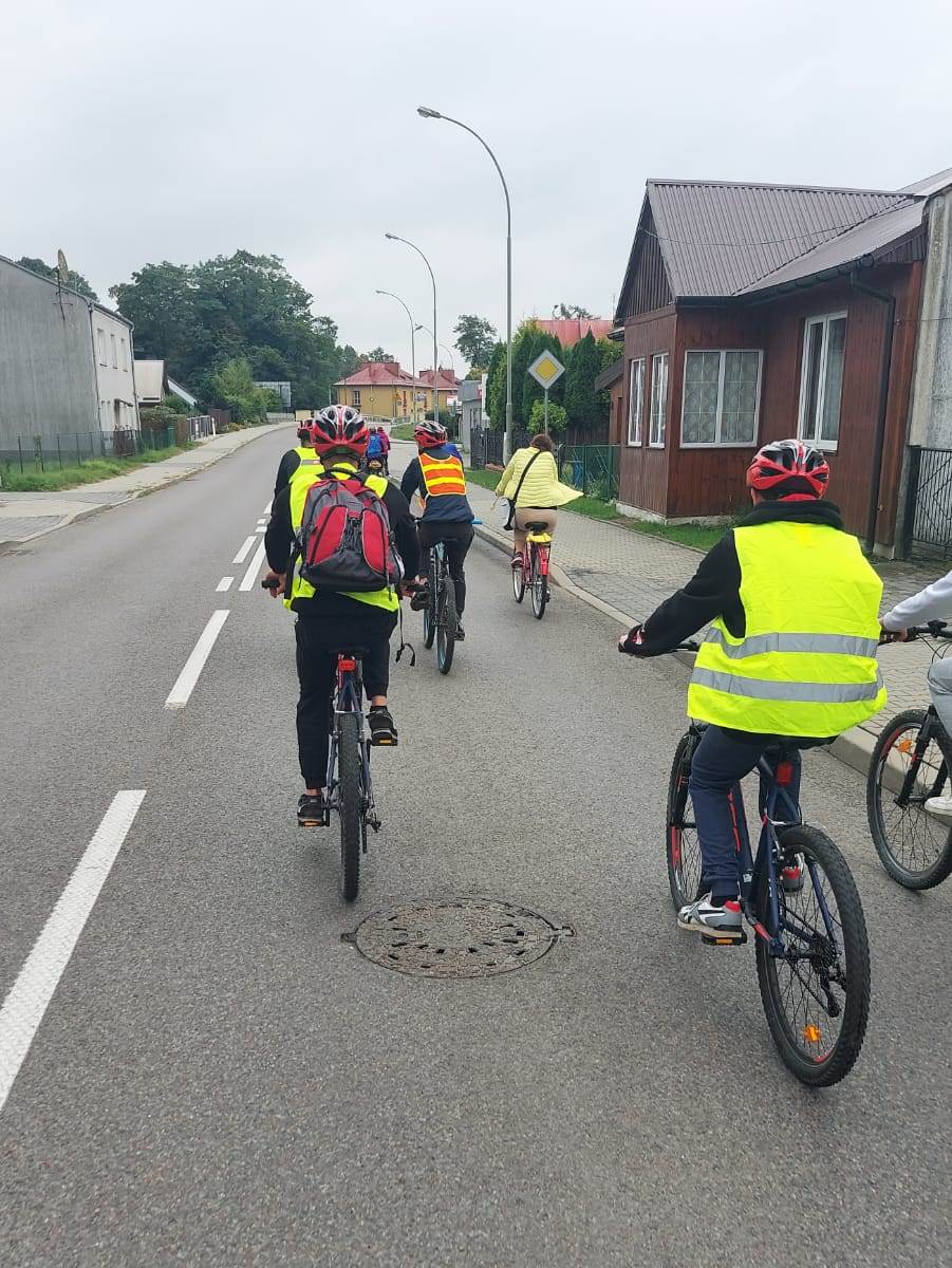 Rajd rowerowy | Wychowankowie MOW w kaskach i kamizelkach odblaskowych widziani od tyłu jadą ulicą na rowerach, po bokach drogi niskie zabudowania..jpg