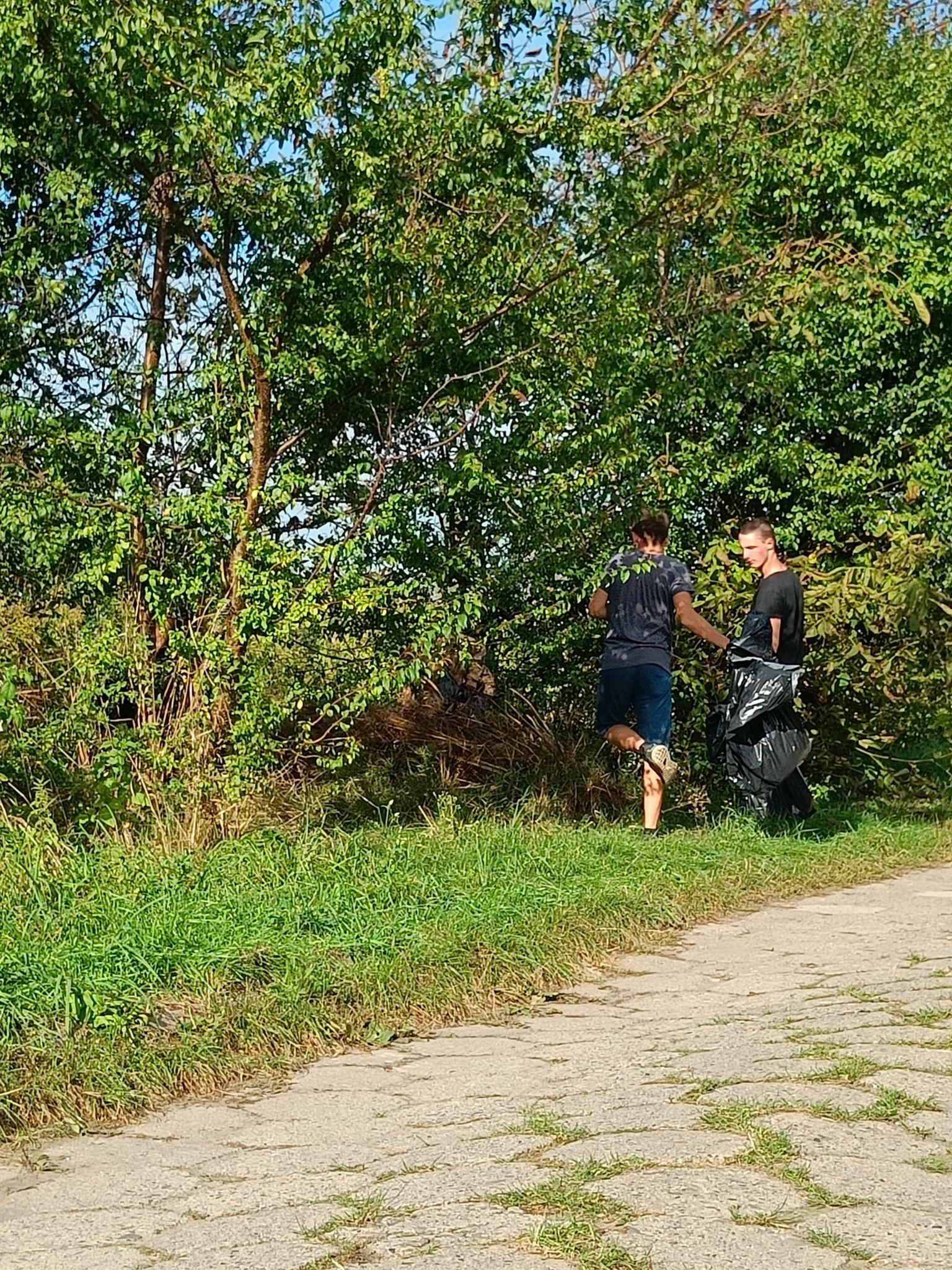 Sprzątanie świata | Dwóch wychowanków MOW stoi tyłem między drzewami, trzymając duży czarny worek, z przodu widać fragment drogi..jpg