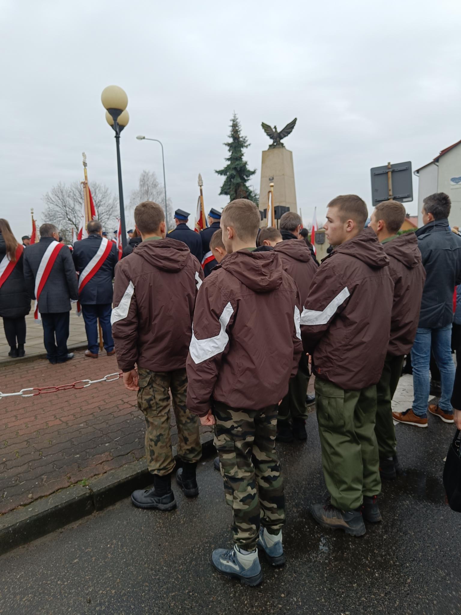 Narodowe Święto Niepodległości | Wychowankowie MOW w brązowych kurtkach i wojskowych spodniach stoją tyłem między innymi ludźmi pod Pomnikiem Niepodległości..jpg