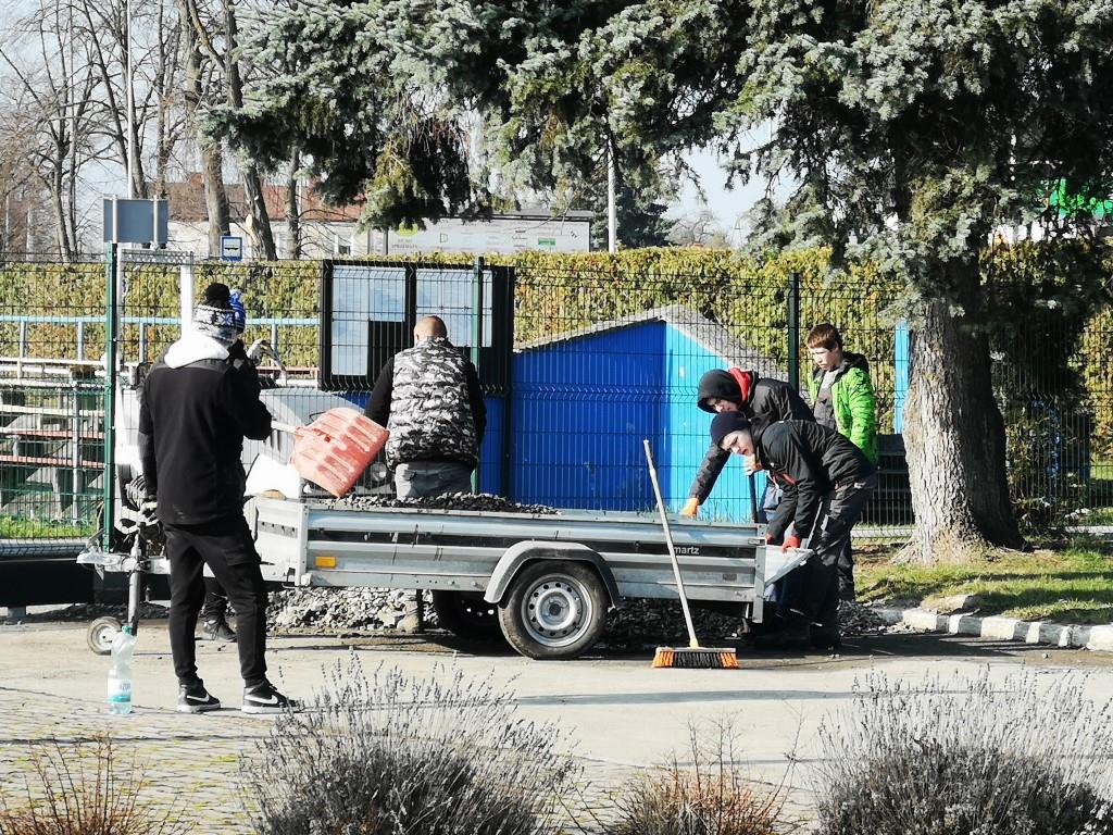 Porządki na stadionie | Kilka osób stoi dokoła przyczepki samochodowej załadowanej gruzem Dwie z nich zapina burtę przyczepki.jpg