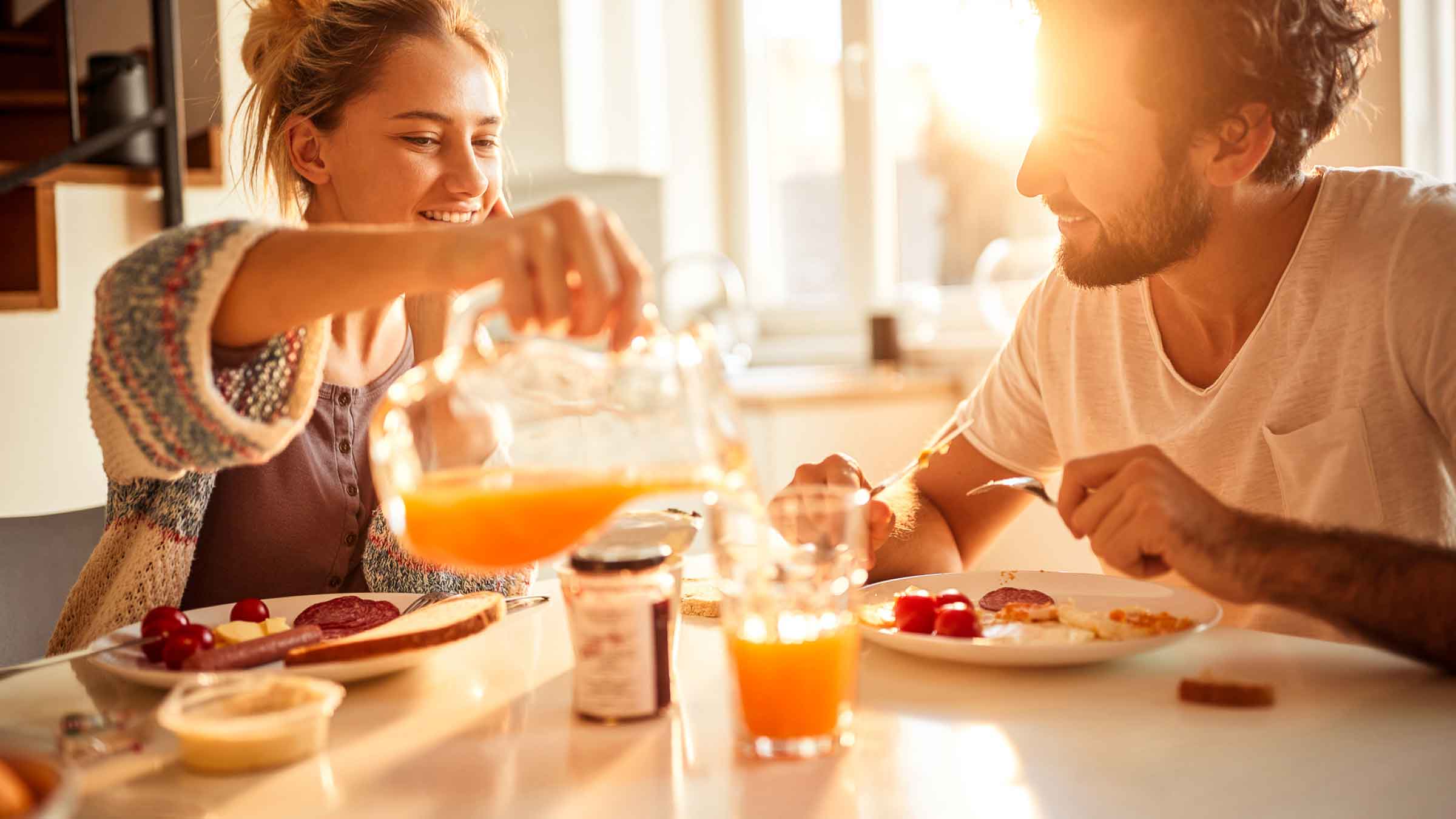 Young couple at breakfast table, the woman is pouring orange juice from a jug into her partners glass. The sun is shining brightly through the large window in the background.