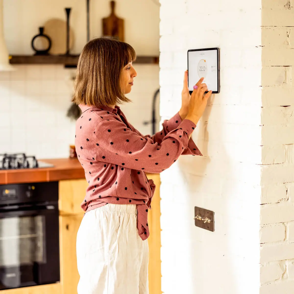 Middle aged woman in pink polka dot shirt, uses touch screen on a tablet mounted to her kitchen wall to adjust the temperature via her ducted air conditioning system.