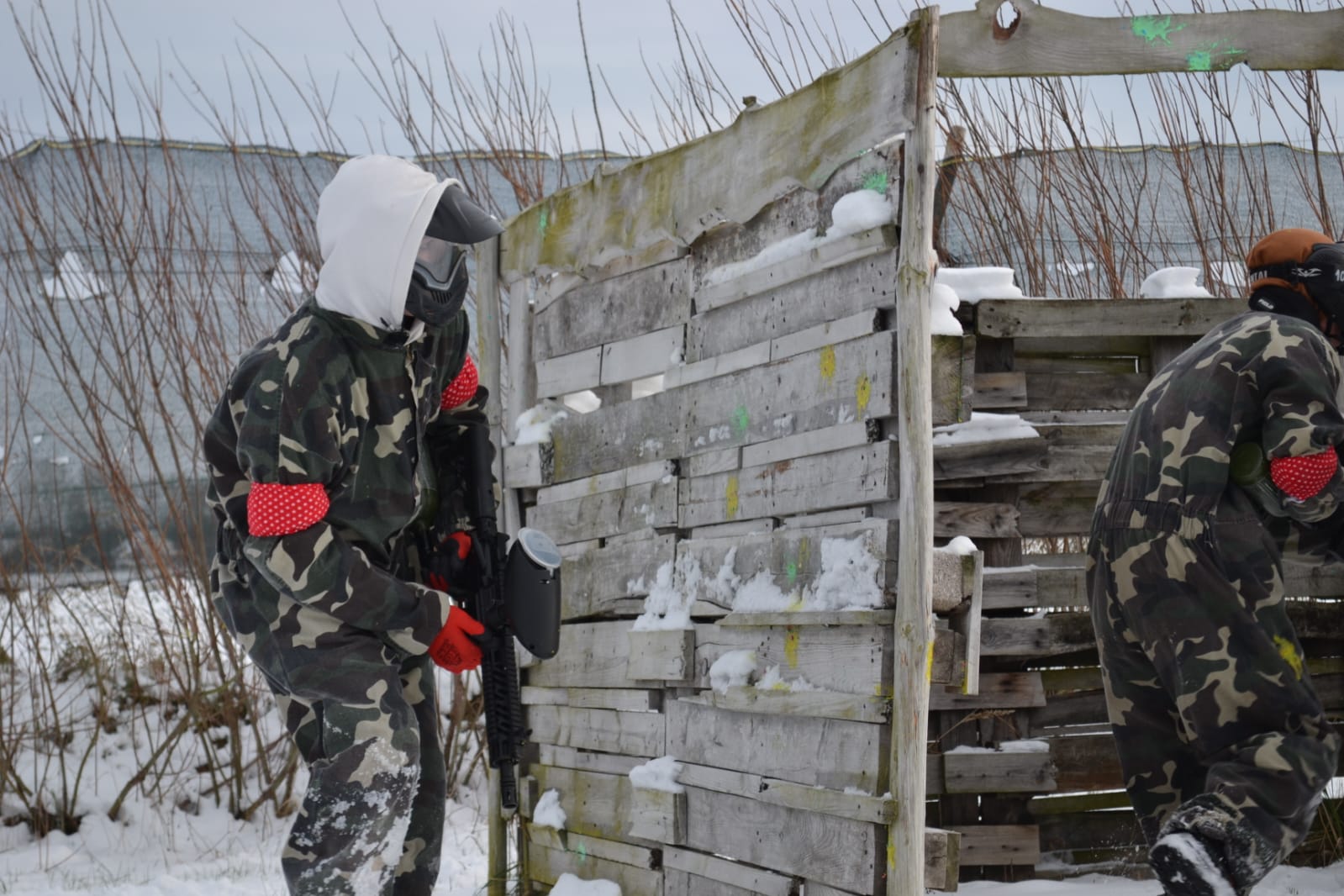 Wycieczka integracyjna klas Branżowej Szkoły I Stopnia w MOW w Lubaczowie | Postać w stroju do paintballa trzyma w ręku karabinek i chowa się za ścianką z desek. Obok podobnie ubrana postać obrócona tyłem.jpg