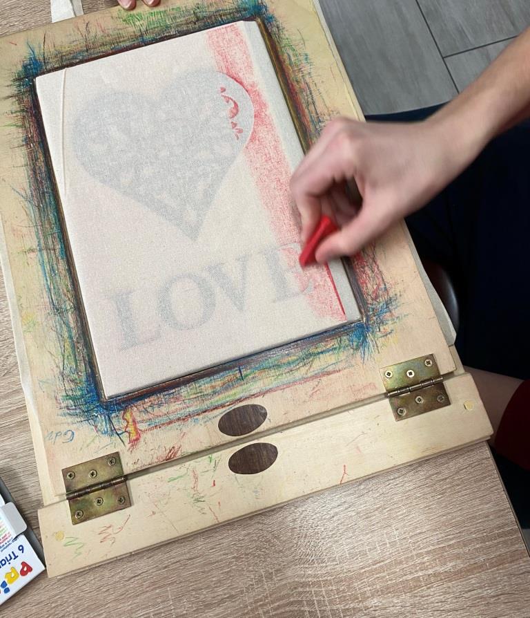 Wizyta w Warsztatach Terapii Zajęciowej w Oleszycach | Dłoń trzymająca czarwoną kredkę woskową rysuje po kartce odbijając widoczne pod spodem serce i napis love.jpeg