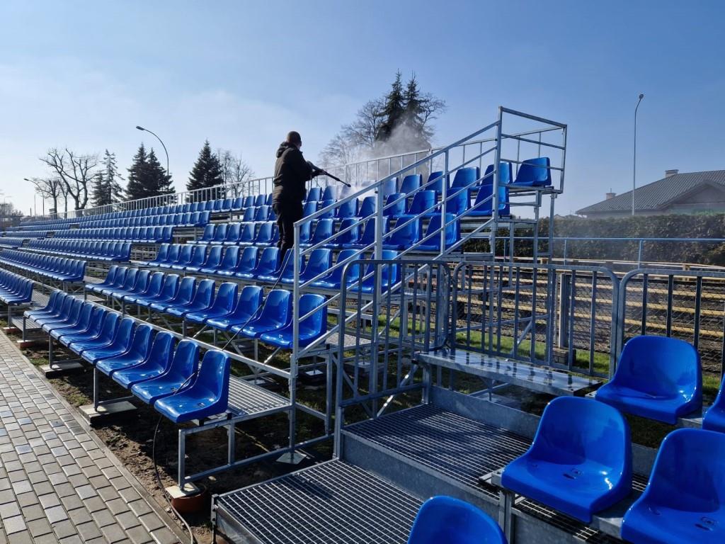 Porządki na stadionie | Ubrany na czarno chłopiec stoi między niebieskimi krzesełkami na trybunach myjąc je myjką ciśnieniową.jpg