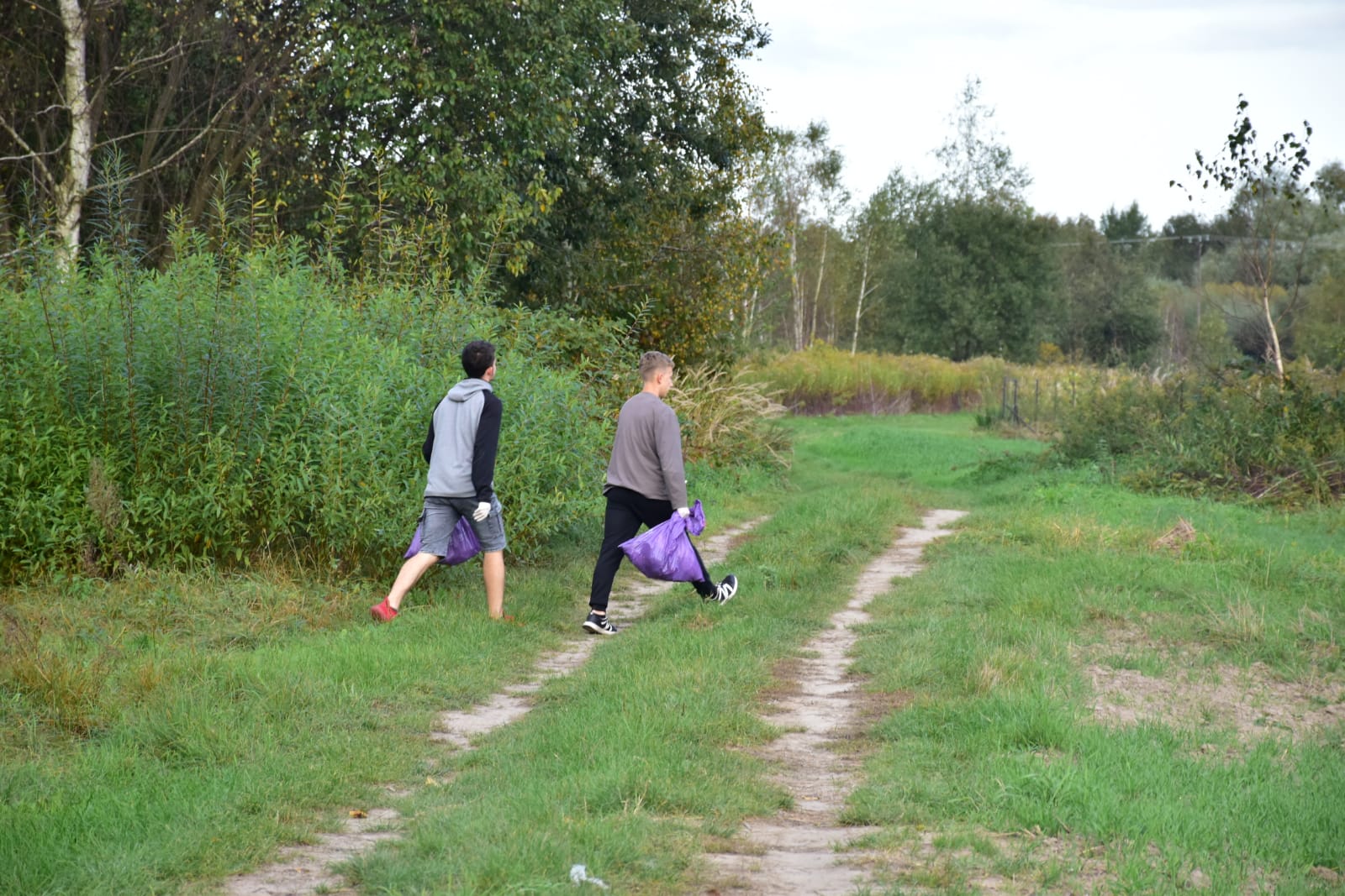 Sprzątamy dla Polski | Dwóch chłopców wychodzi z lasu na leśną drogę. Obaj niosą fioletowe worki. W głębi zdjęcia las, na pierwszym planie trawa..jpg