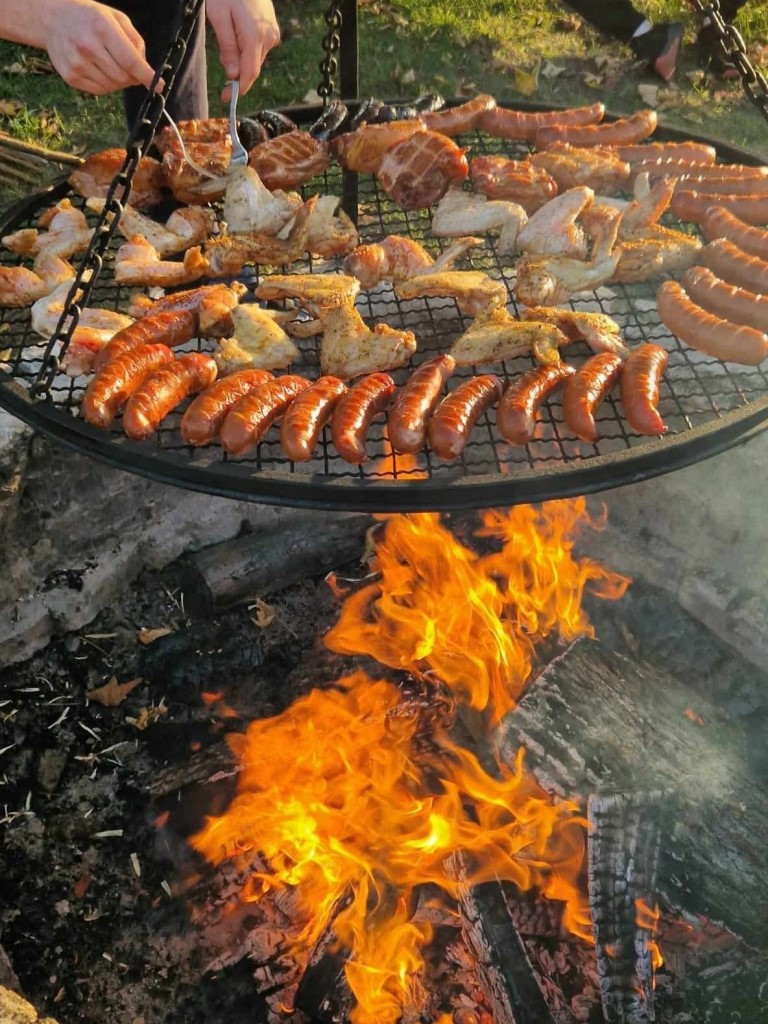 Obóz w Hucie Różanieckiej | Palące się ognisko, nad którym zawieszony jest ruszt z piekącymi się kiełbaskami i mięsem z kurczaka. W rogu zdjęcia widoczne dłonie, które widelcami obracają piekące się mięso..jpg