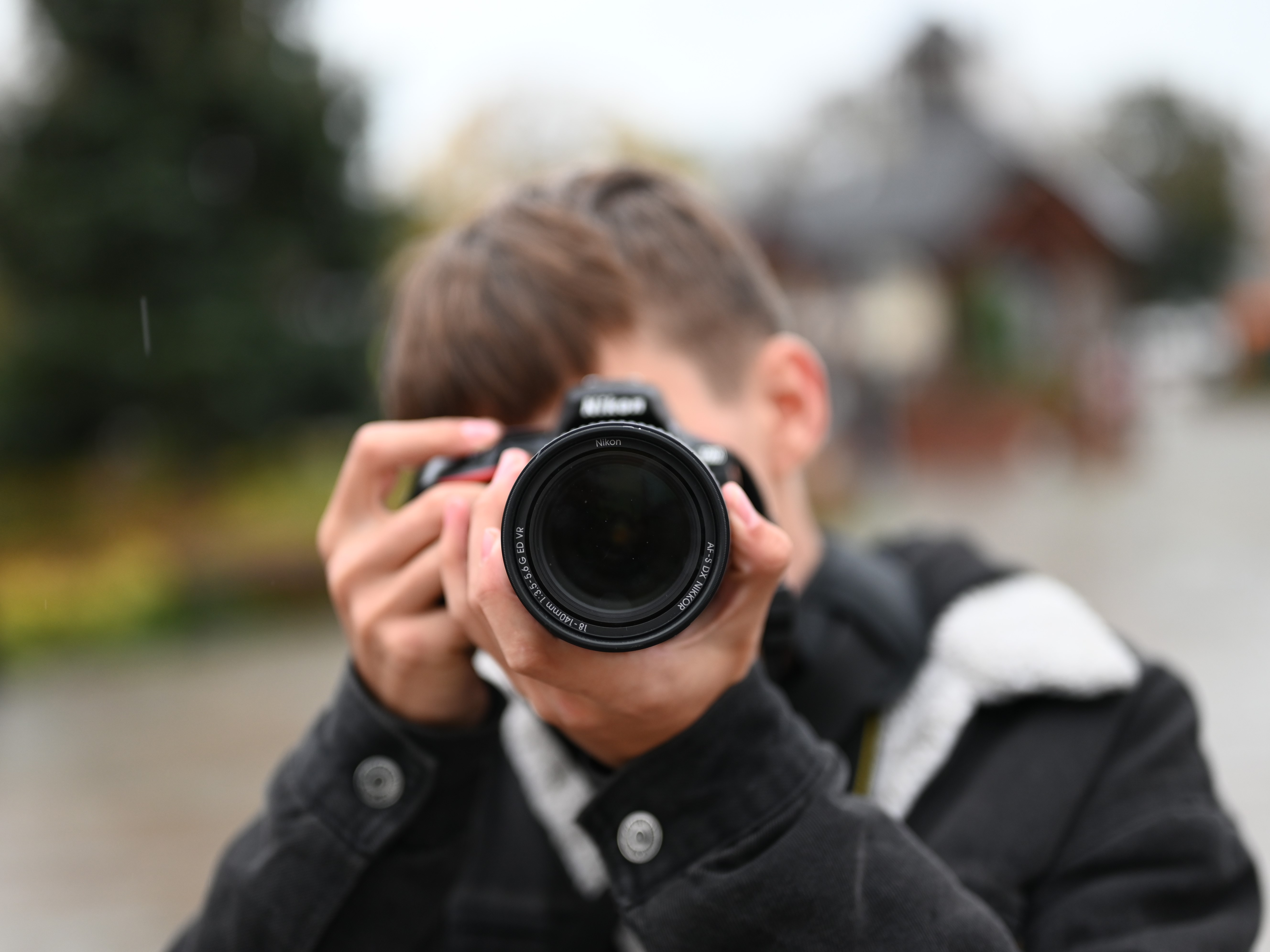 Fotografia jeszcze raz | Chłopiec w ciemnej kurtce z przyłożonym do twarzy aparatem robiący zdjęcie fotografującemu. W tle rozmazane kontury studni na Rynku oraz drzew..jpg