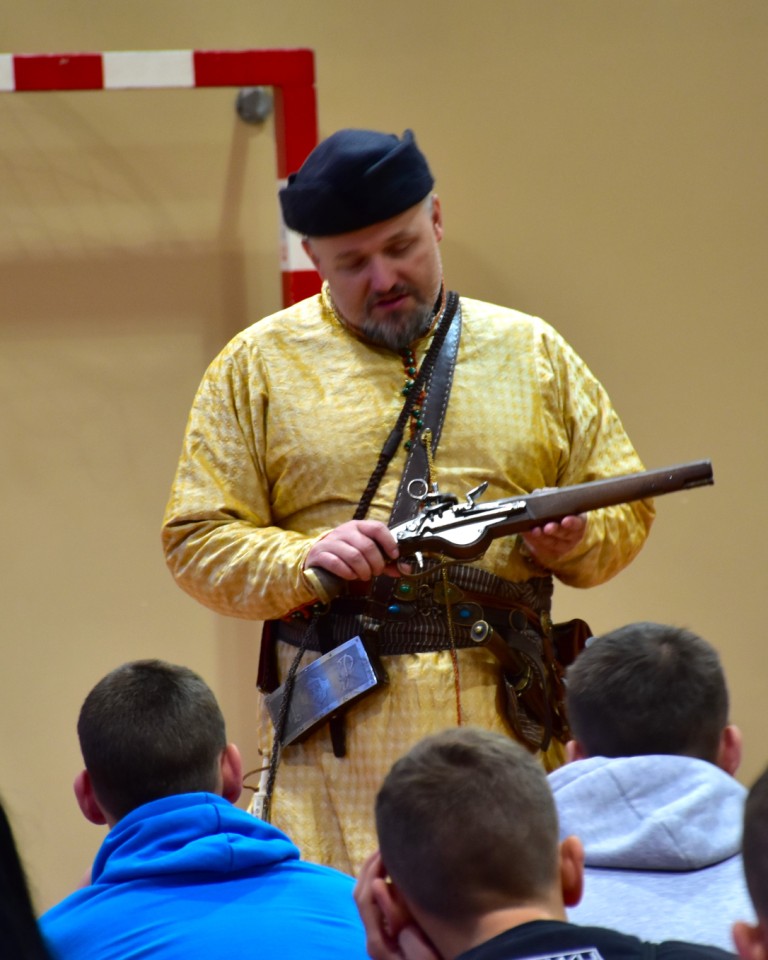 Lekcja historii | Mężczyzna w złotym stroju szlacheckim z ozdobnym pasem prezentuje siedzącym przed nim wychowankom broń palną - duży pistolet..JPG